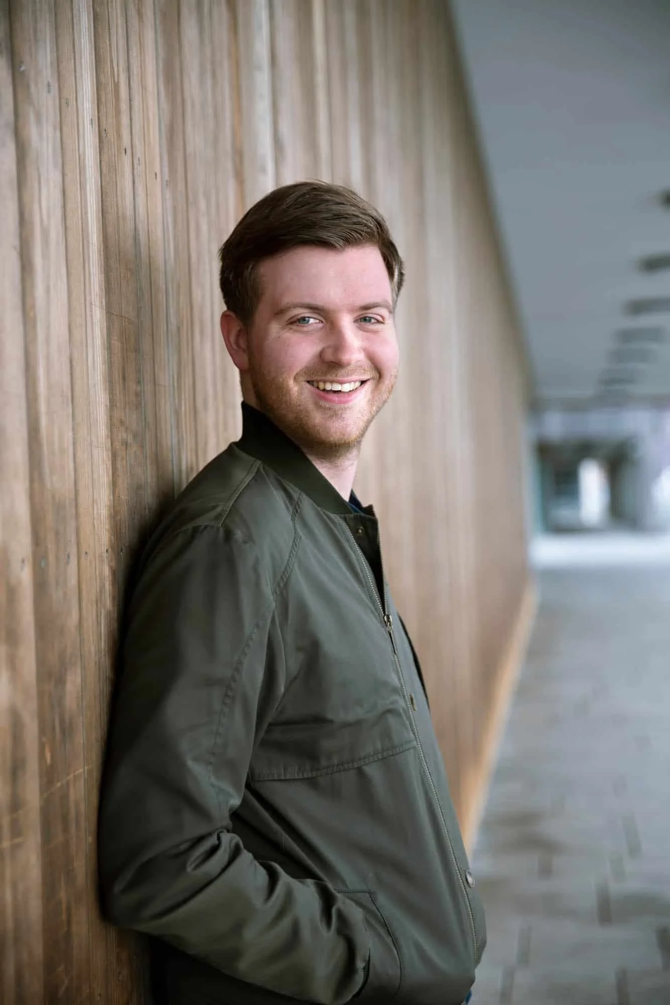 Smilende mann ikledd grønn bomberjakke lener seg mot en trevegg i en lang gang