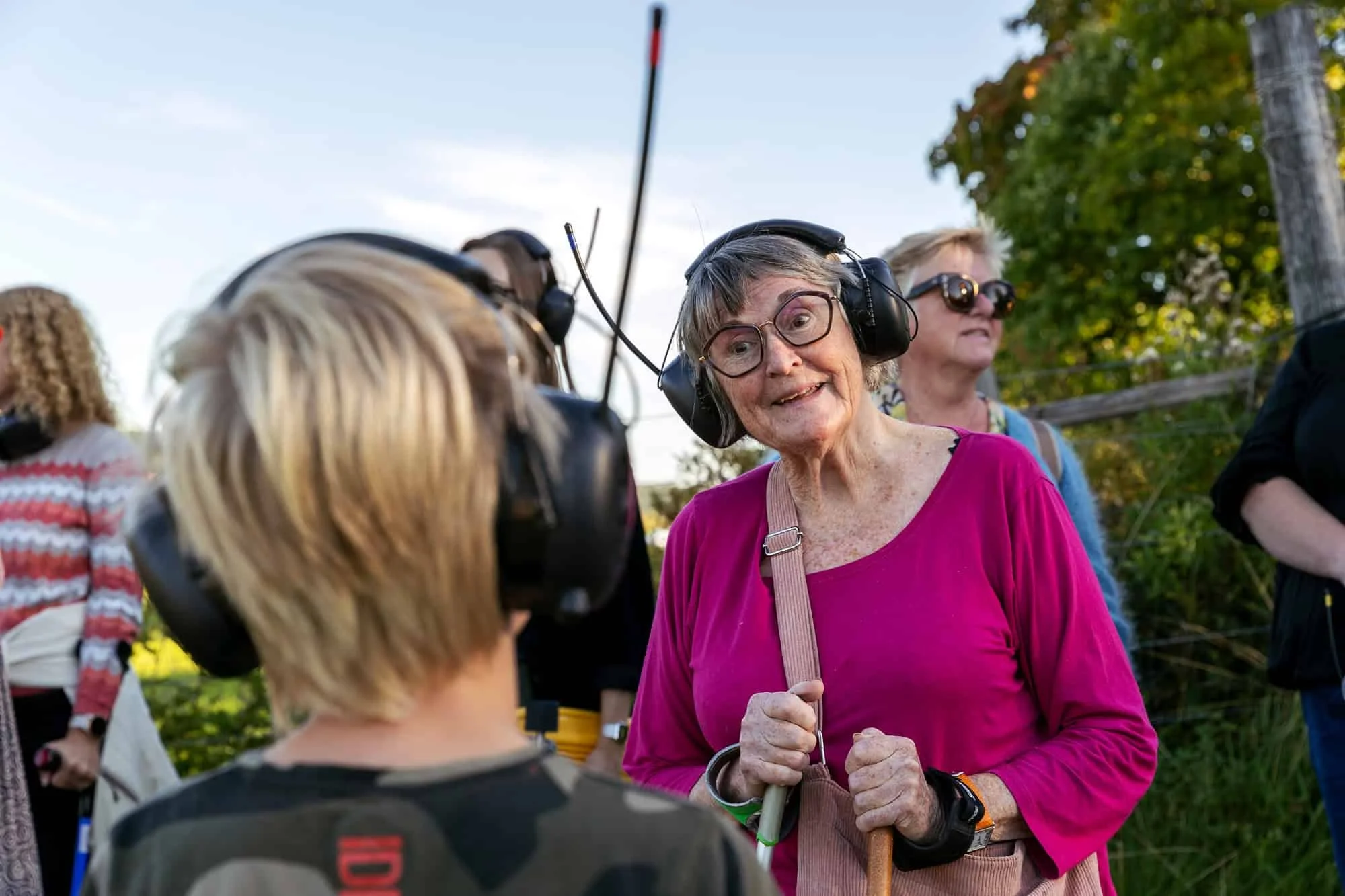 Eldre kvinne kledd i rosa genser med høretelefoner på hodet står sammen med andre mennesker med høretelefoner