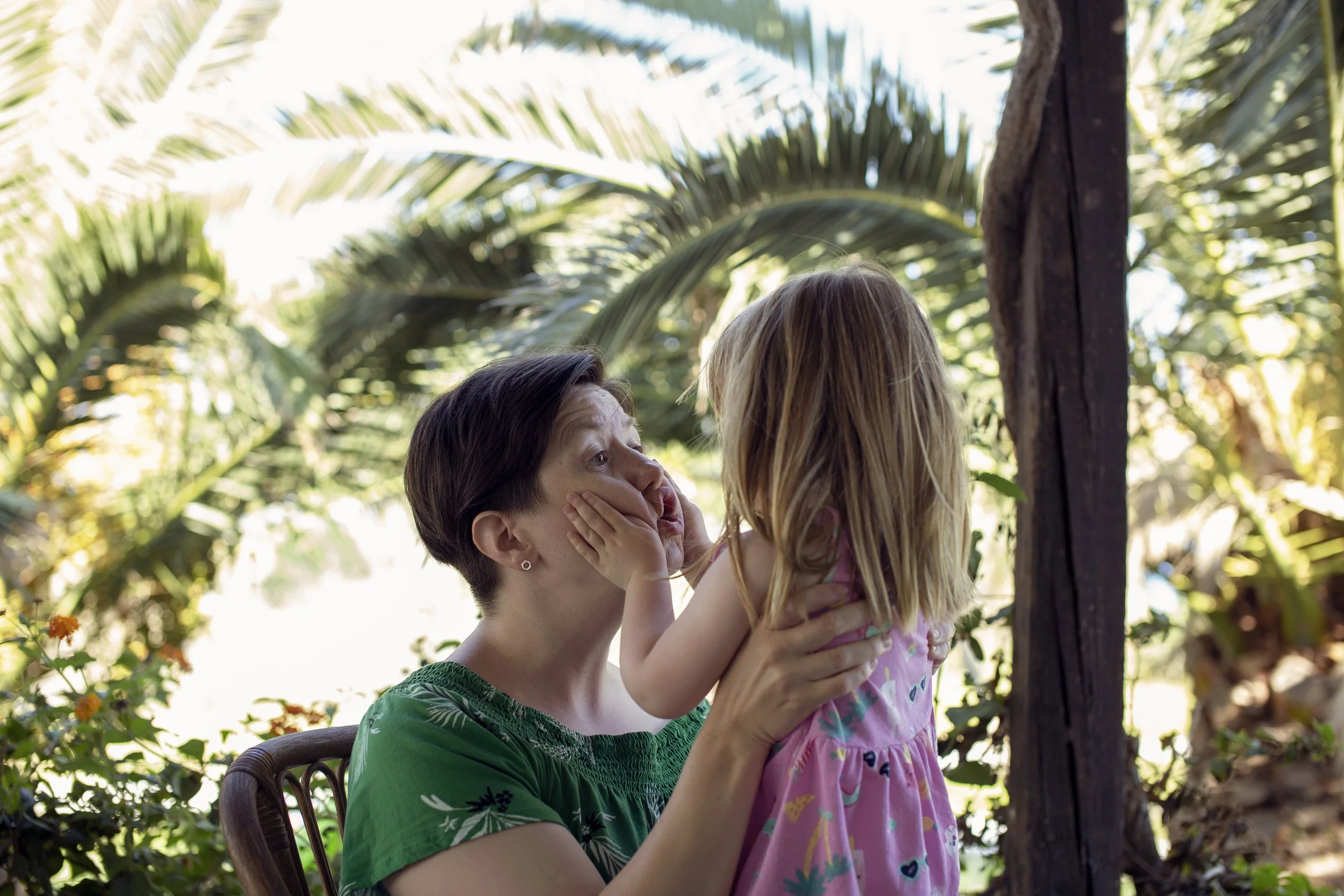 Liten jente i rosa kjole lager grimaser på moren sin med palmer i bakgrunnen