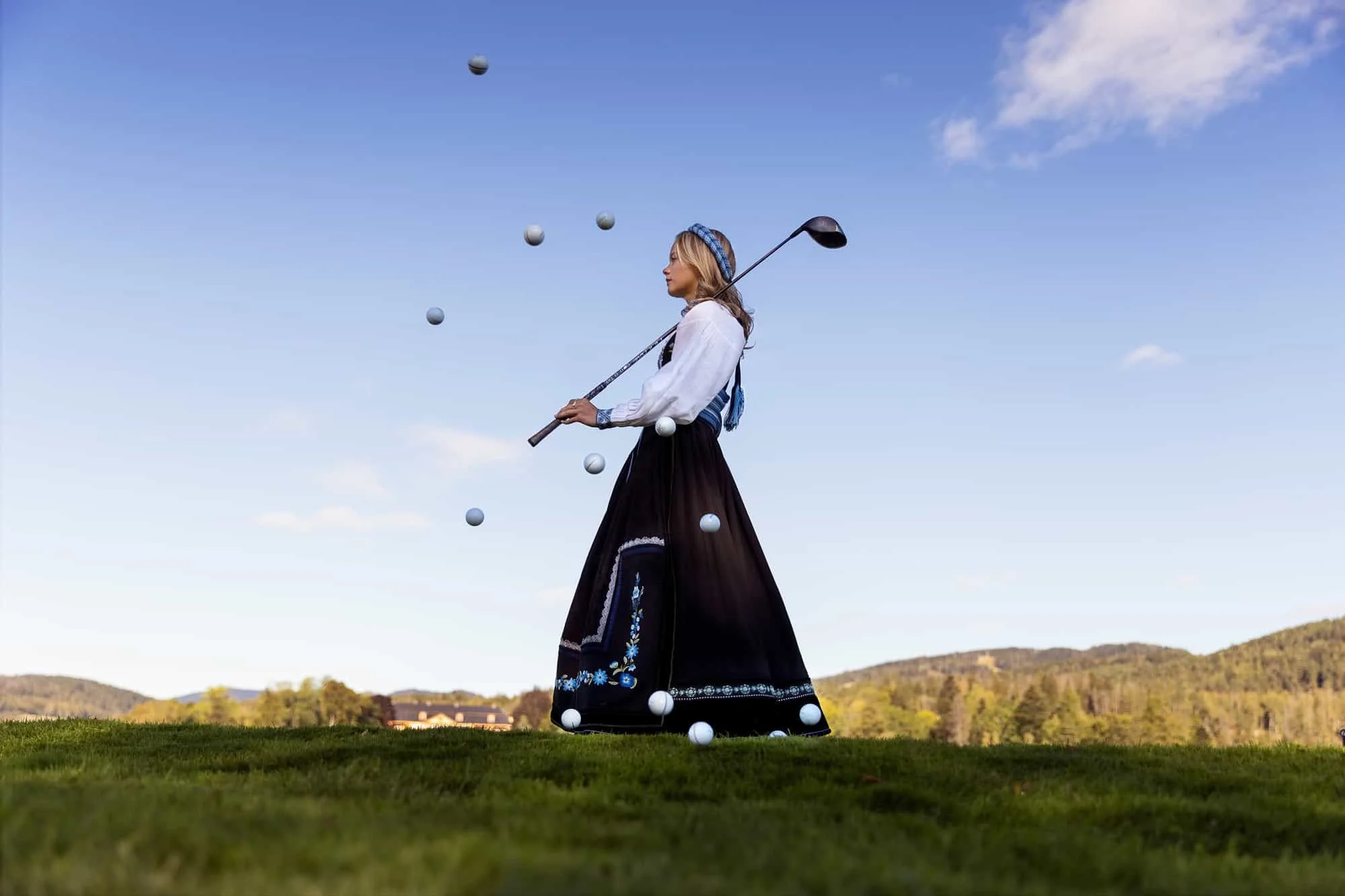 Konfirmant i bunad i profil med golfkølle på skuldra og mange golfballer i lufta rundt henne