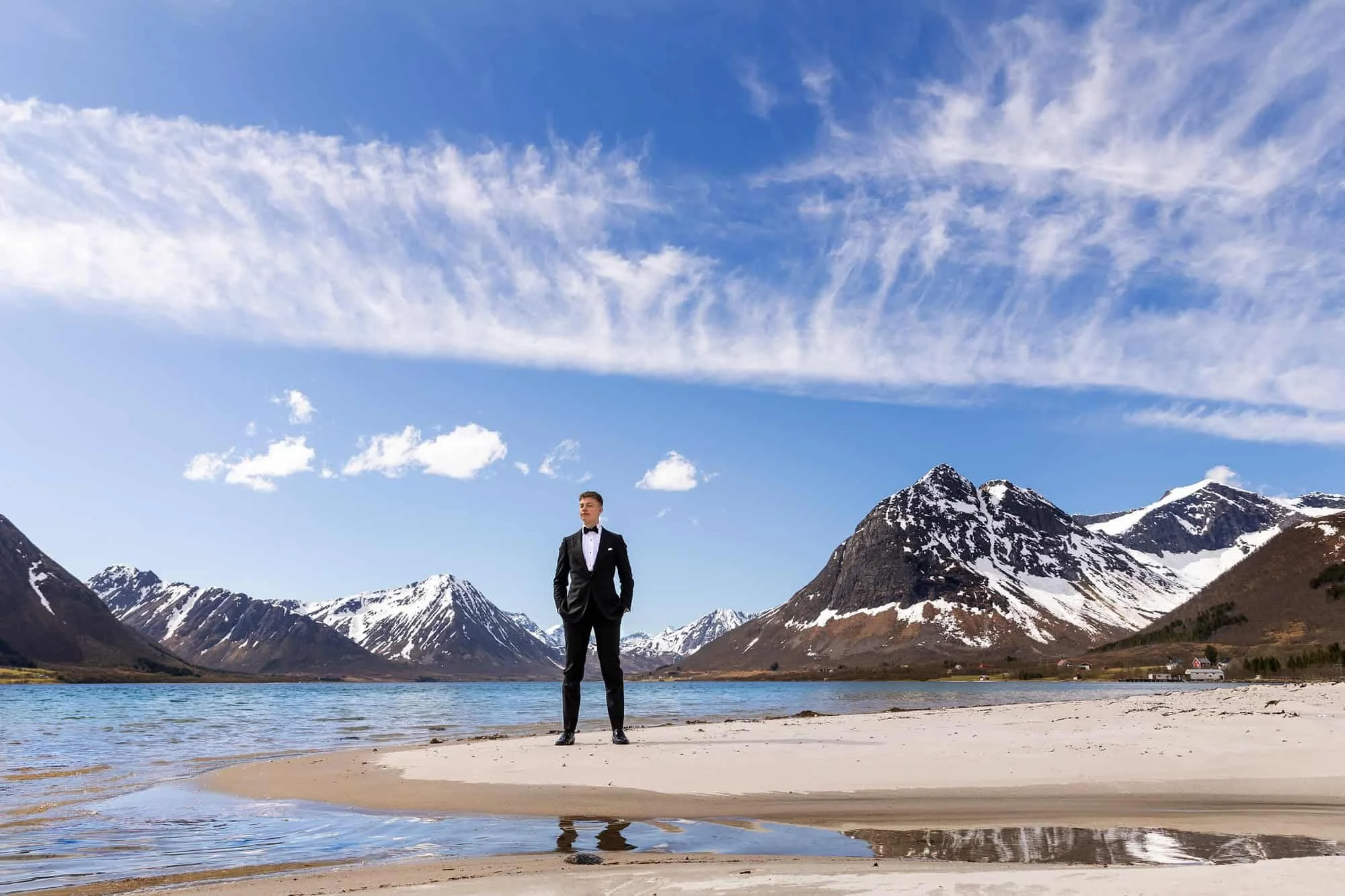 Dresskledd ungdom står på ei hvit sandstrand blant snøkledde fjell og blått hav