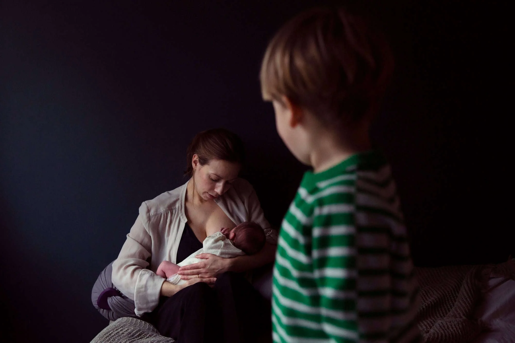 En mor ammer barnet sitt i et sort rom mens storebroren står i forgrunn i stripete genser og ser på