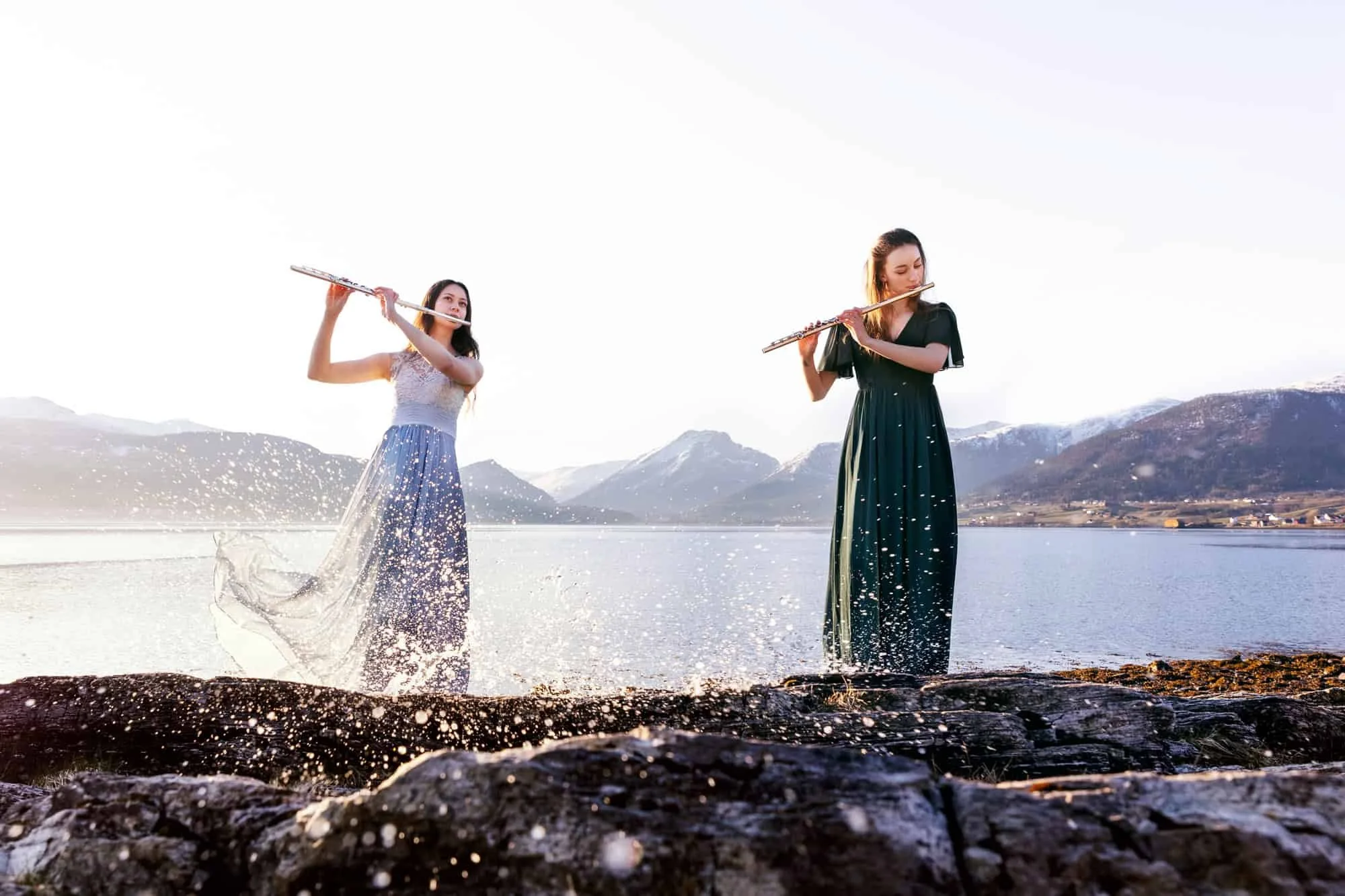 To fløytister ikledd lange kjoler står ved havet og spiller mens vann spruter rundt dem