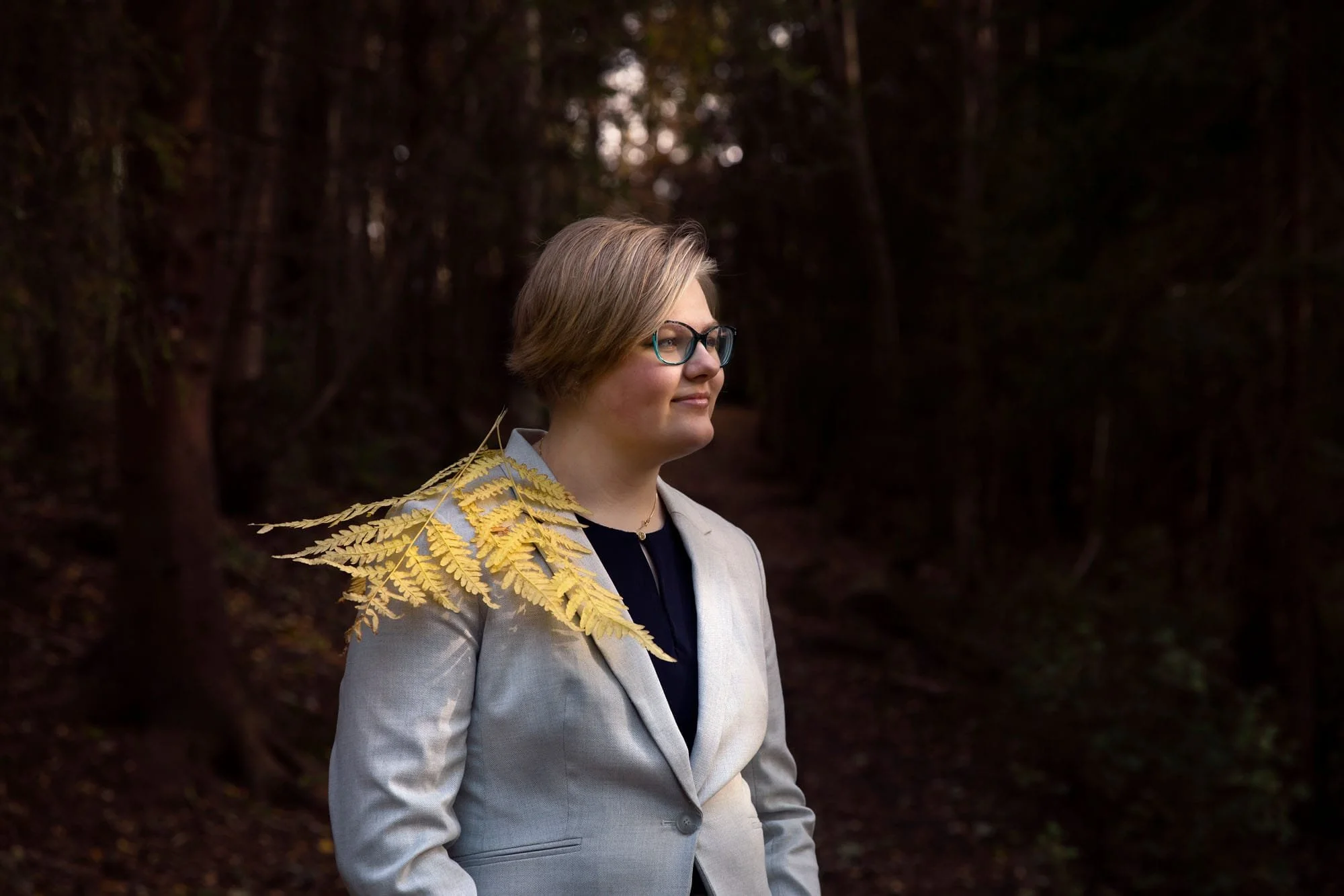Jentekonfirmant med briller og kort hår kledd i lys dress med et stort gult bregneblad på skulderen sin