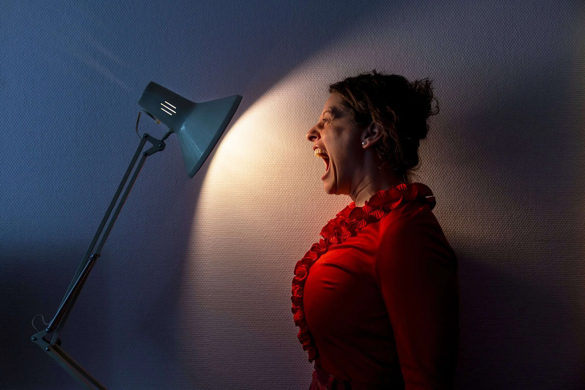 Skuespiller i rød bluse roper mot lyskjegle fra arkitektlampe i mørkt rom på Oppsal Samfunnshus i Oslo.