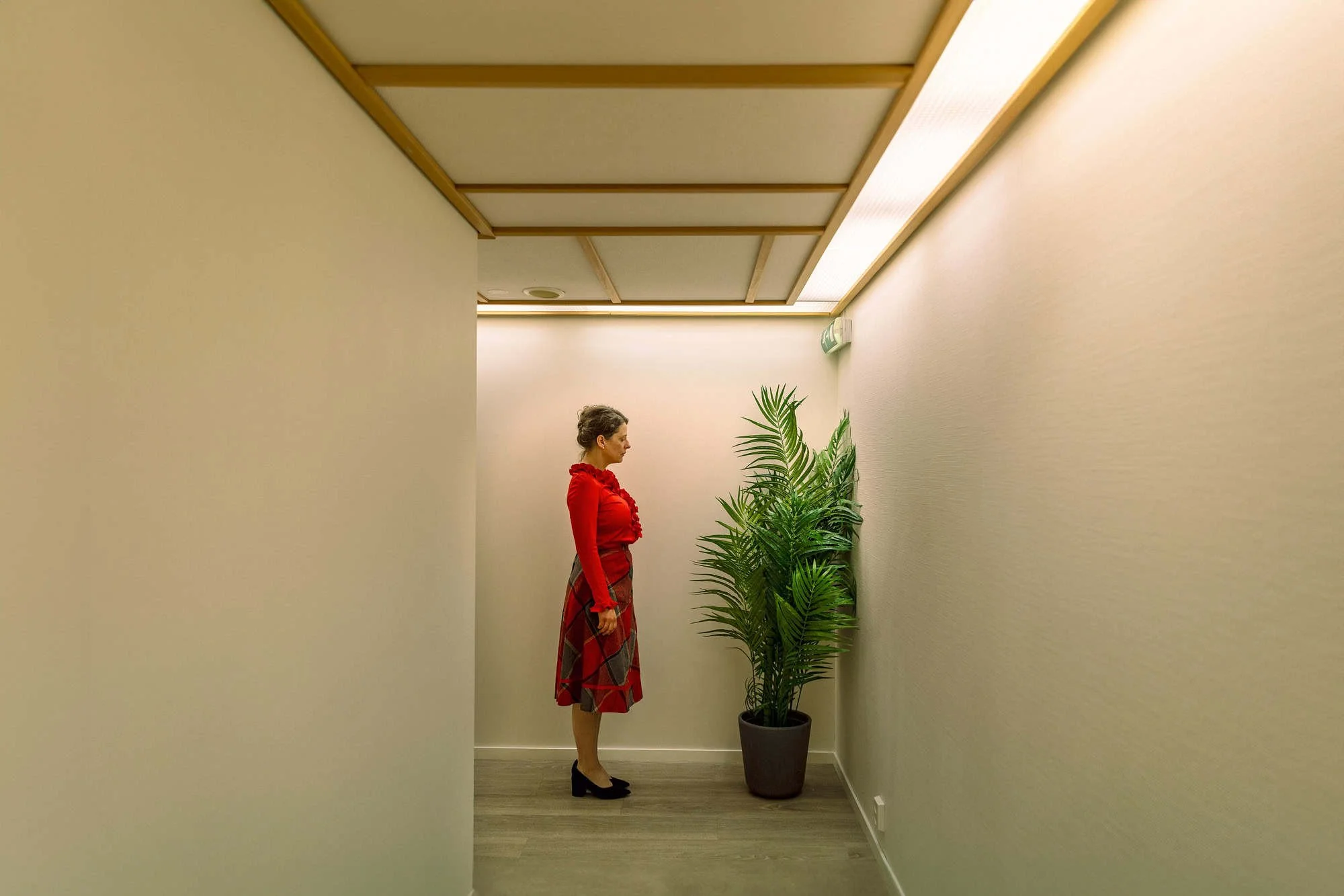 Skuespiller i rødt antrekk står alene i smal korridor med plante på Oppsal Samfunnshus i Oslo.