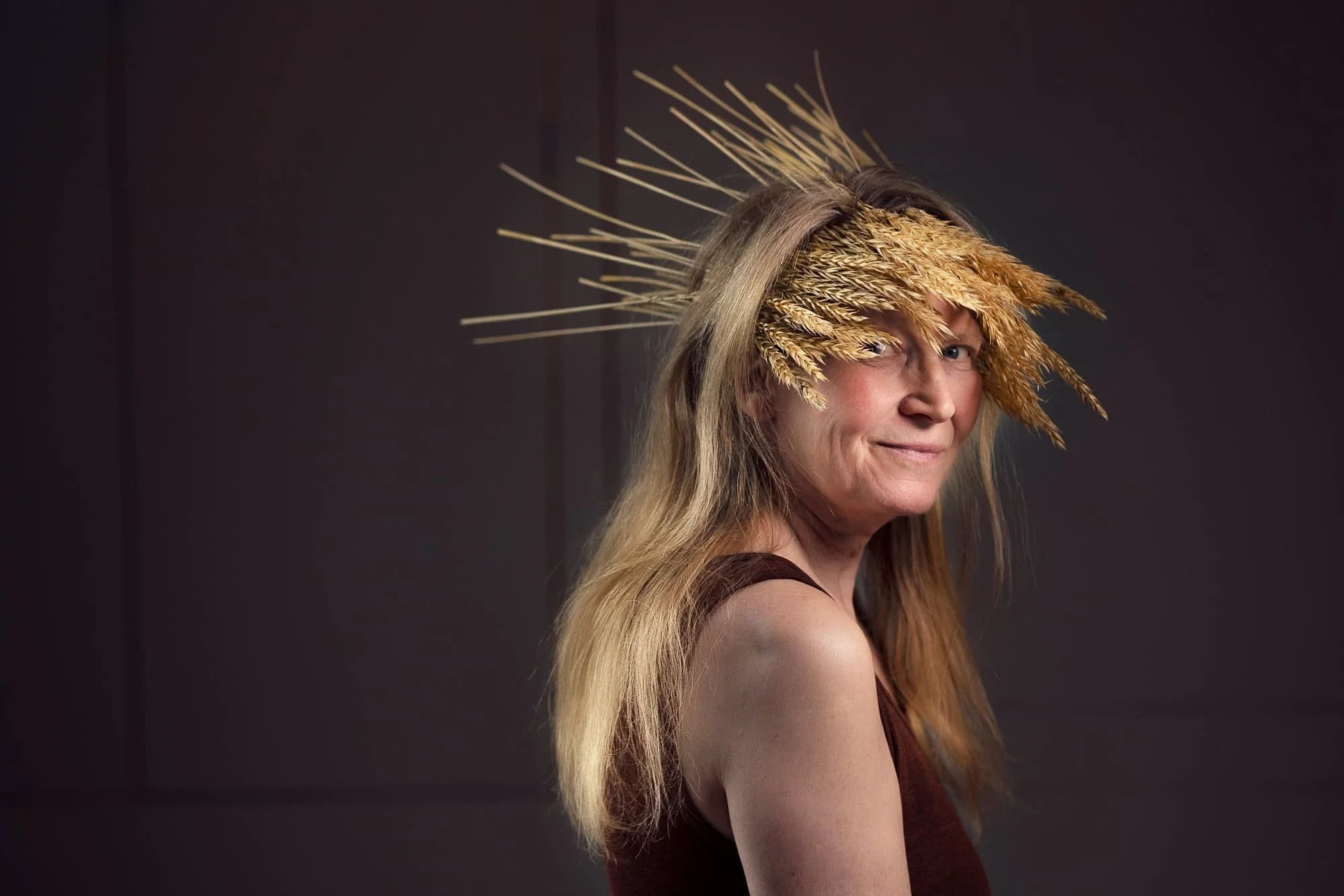 Kunstnerisk portrett av eldre kvinne med strå på hodet som ser rett i kameraet