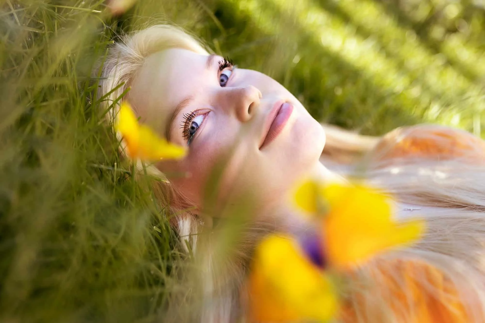 Portrett av ung, lyshåret kvinne som smiler lurt, liggende i en blomstereng med orange bluse