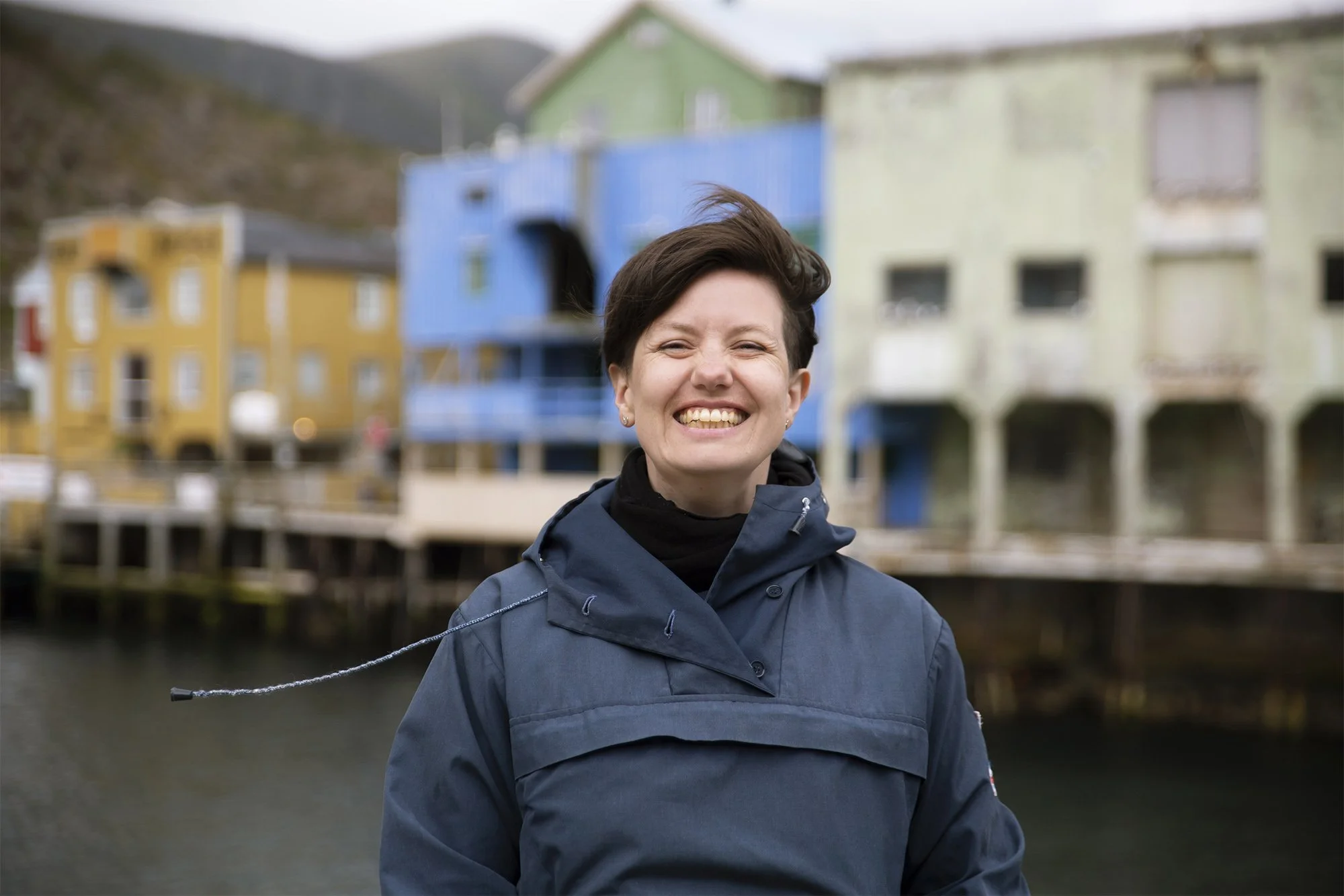 Smilende fotograf i klassisk, blå anolrakk med fargerike bygninger i Nyksund som bakteppe