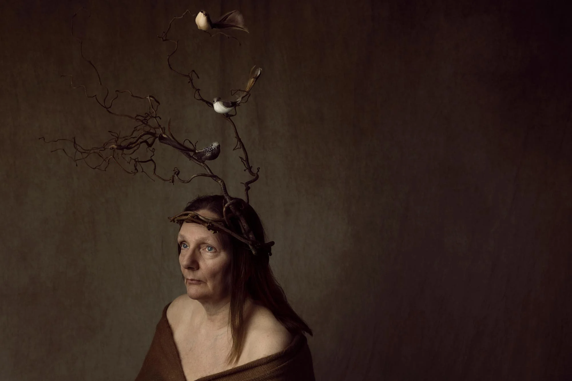 Kunstnerisk studioportrett av eldre kvinne med kvistkrone og fugler på hodet