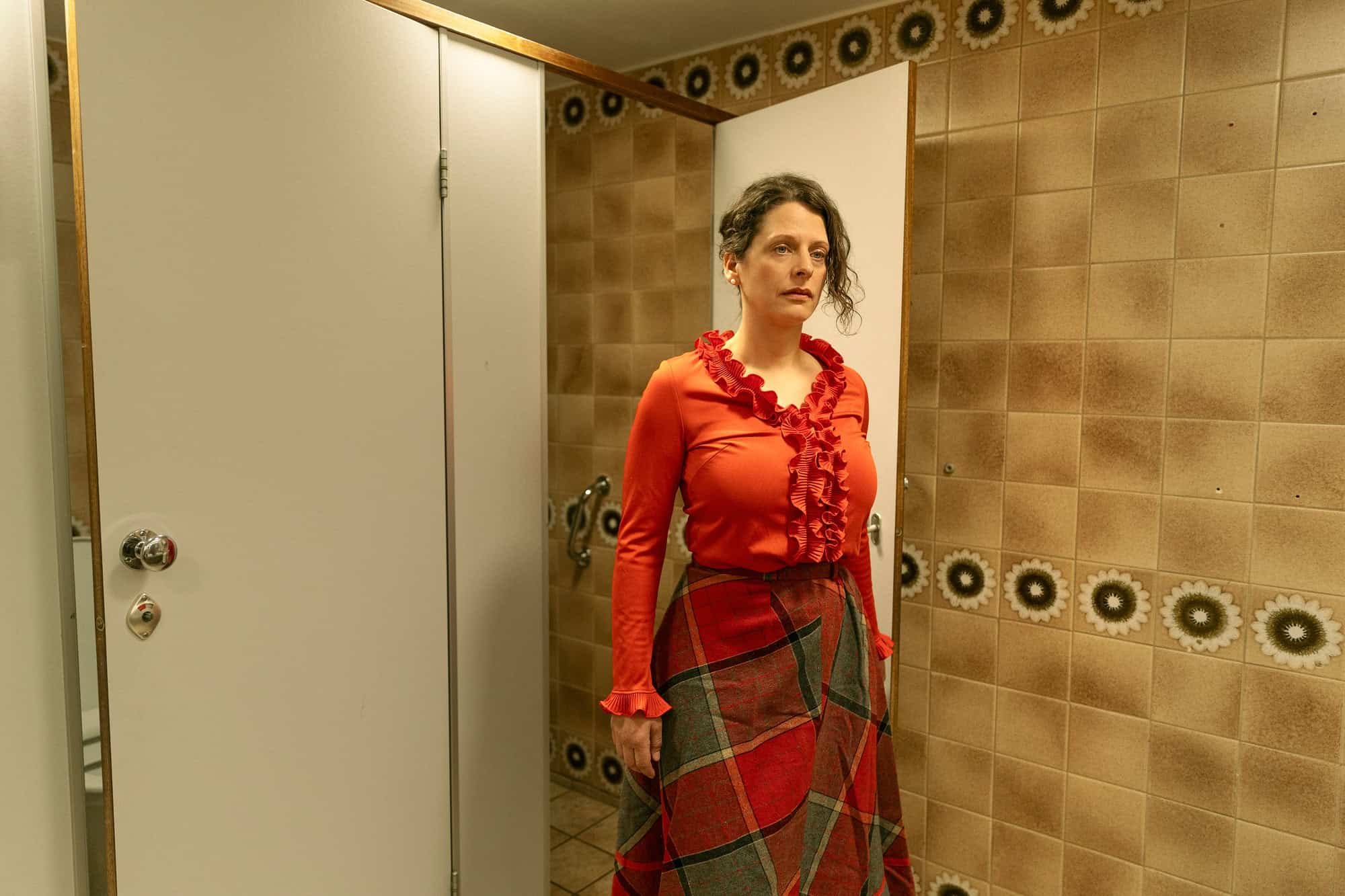 Portrett av skuespiller i rødt antrekk stående i bad med brune fliser og åpen dør.