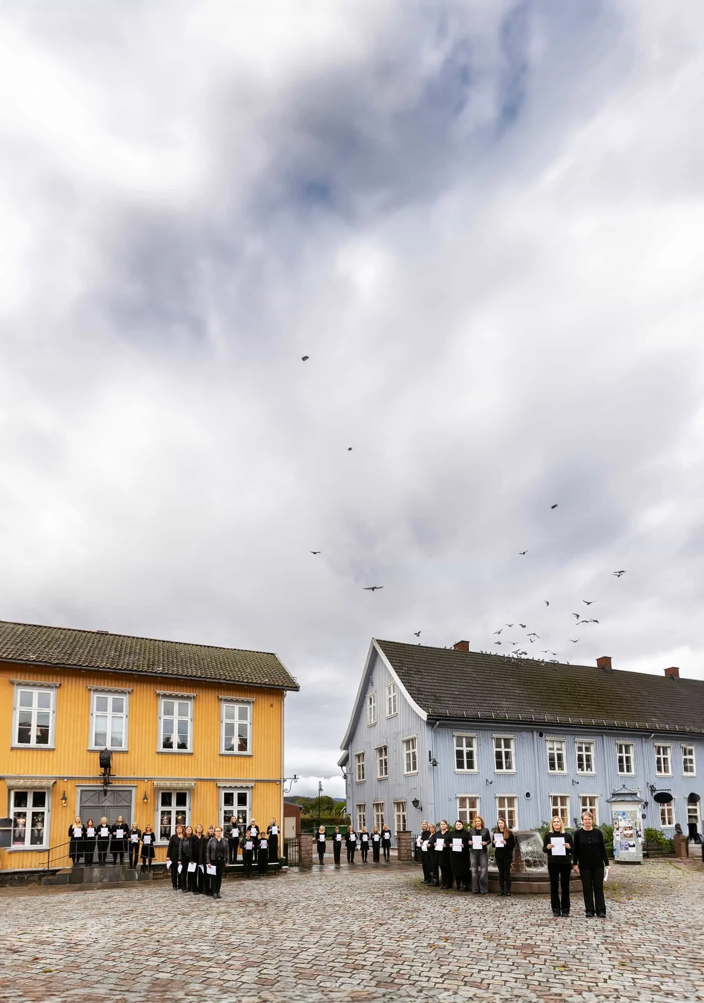 Et sortkledd damekor holder notater foran seg på en åpen plass med fargerike trehus over husene flyr det fugler