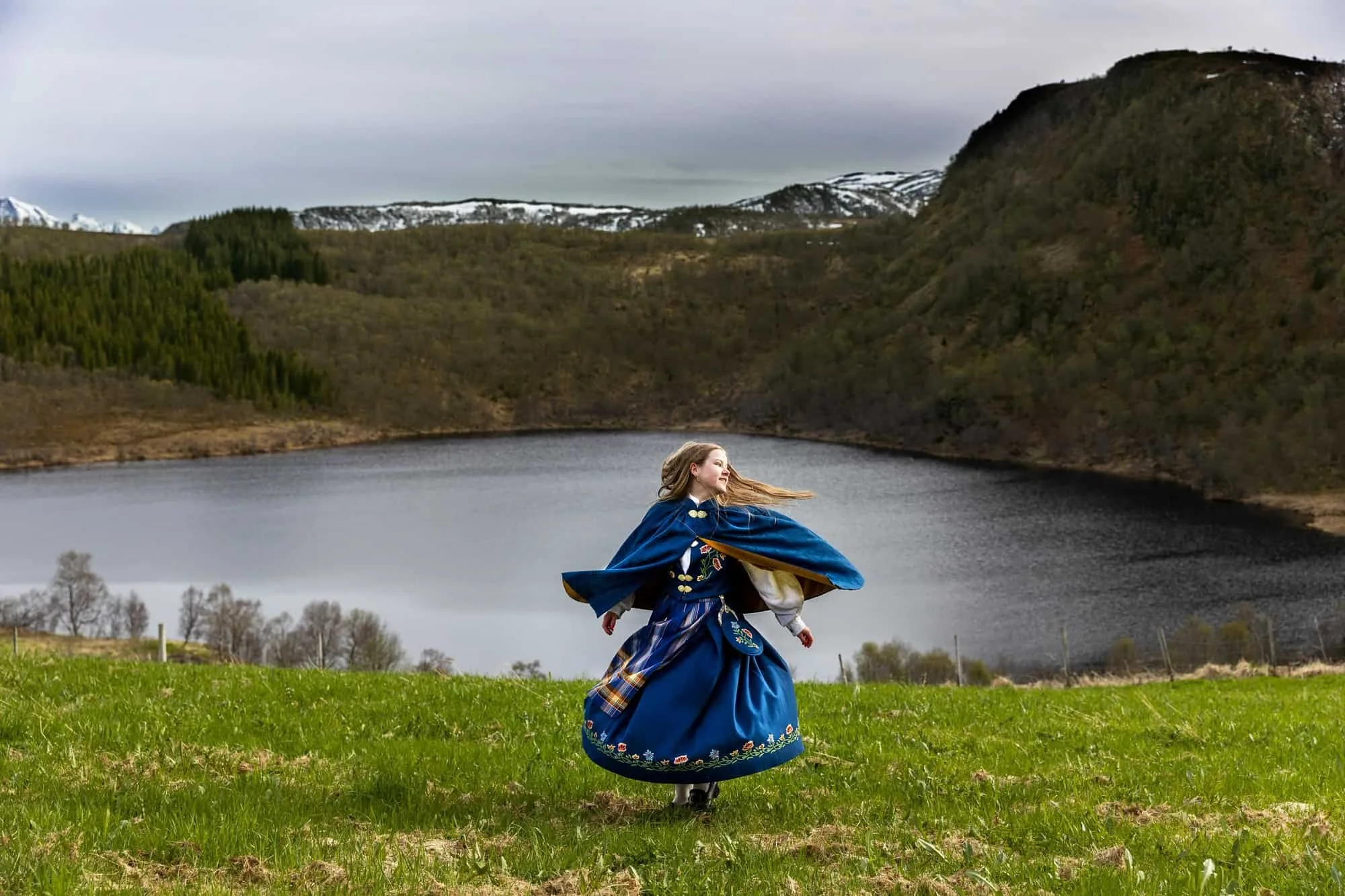 Jente i blå Nordlandsbunad svinger seg rundt i et landskap av vann og fjell