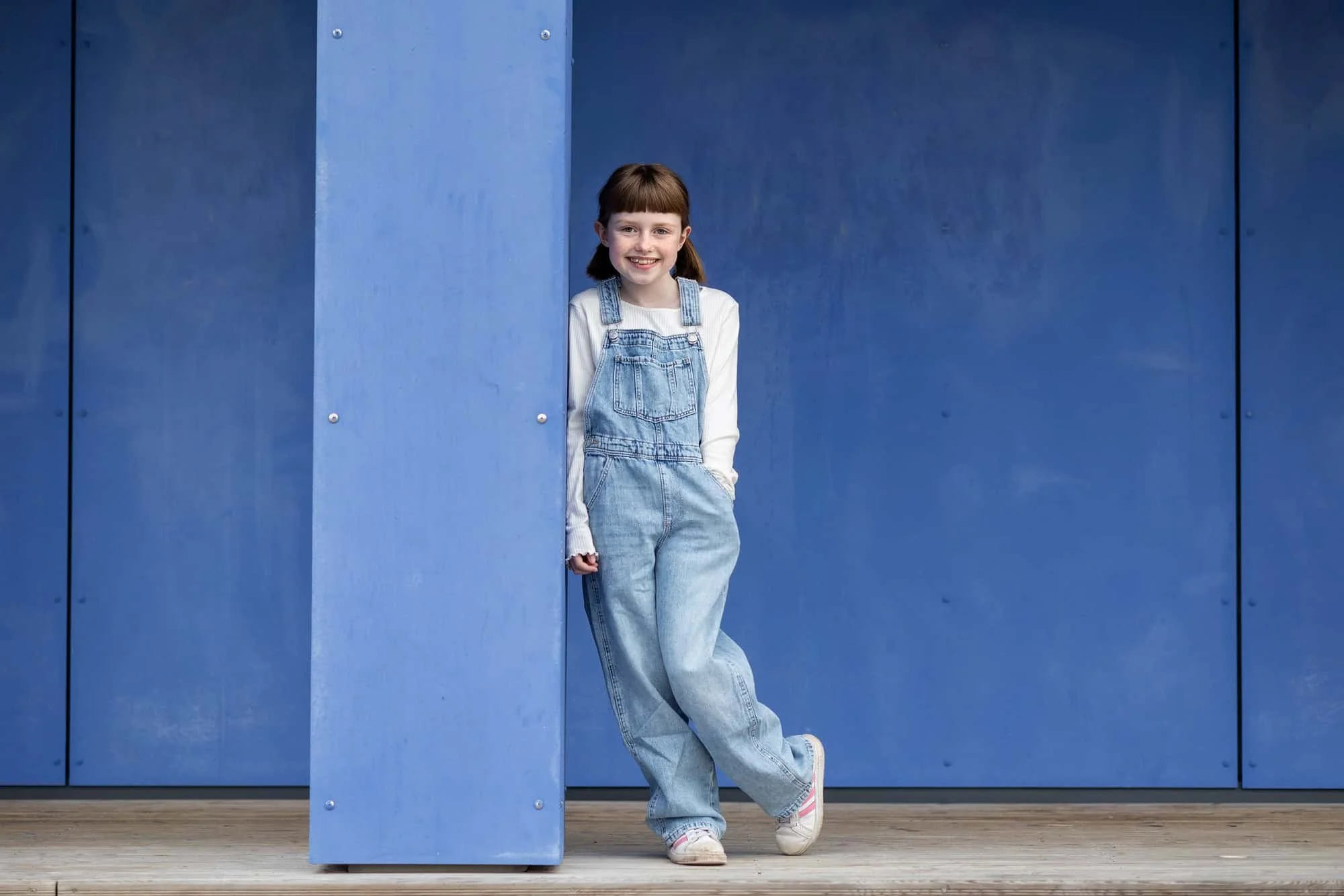 Jente i blå snekkerbukse lener seg mot en blå stolpe mot en mørkeblå murvegg