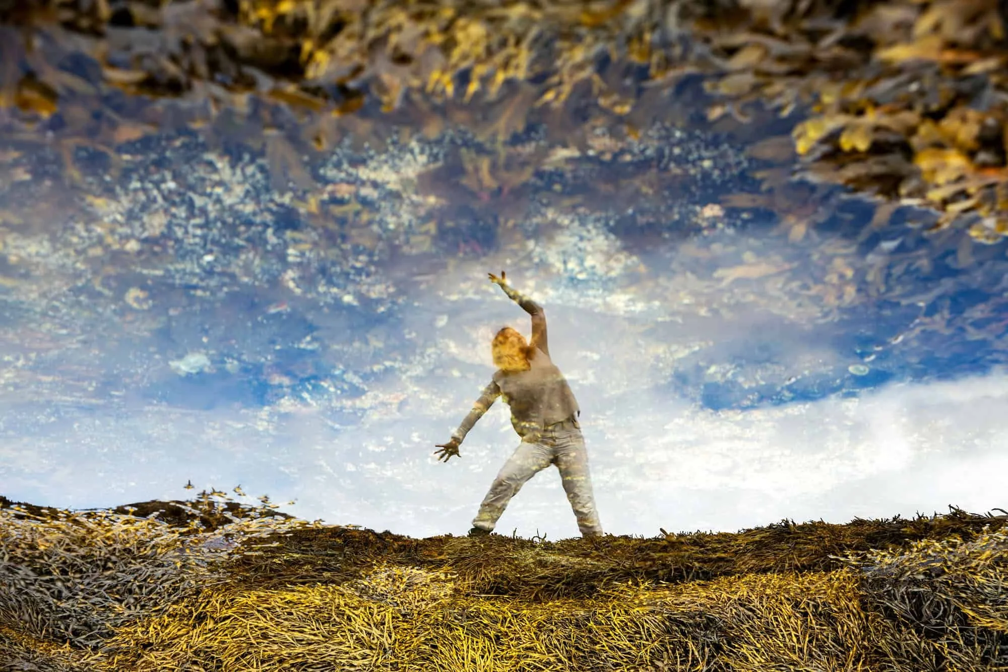 Dame som gjør yoga speiles i vann omgitt av gul tang