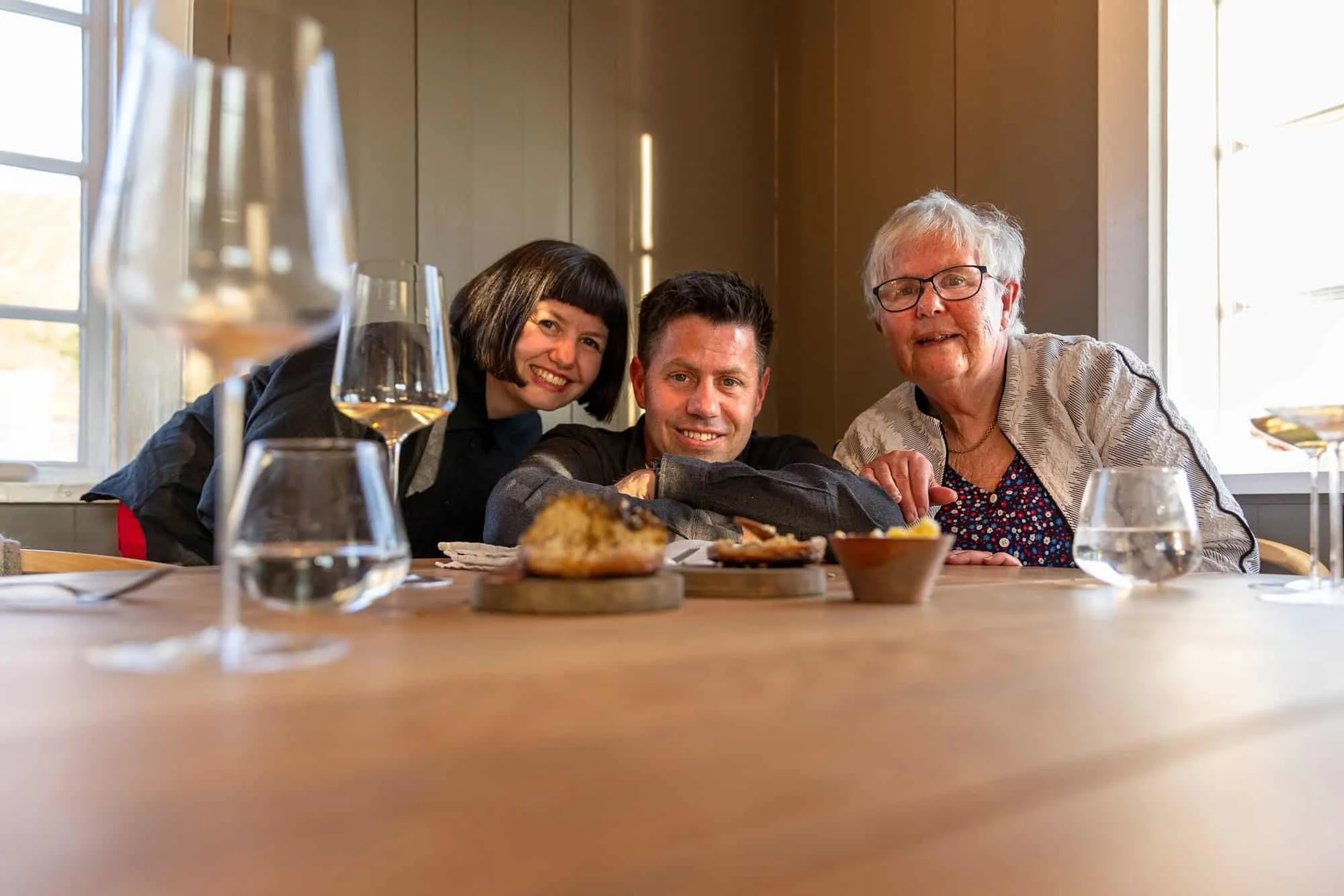 En eldre mor sammen med sine to barn ser i kameraet og smiler over fat med mat på en restaurant