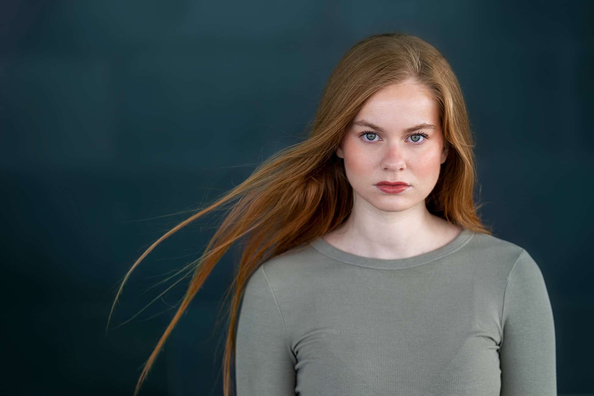 Portrett av kvinne som ser rett i kameraet med rødt hår som flagrer i vinden fotografert mot en sort vegg