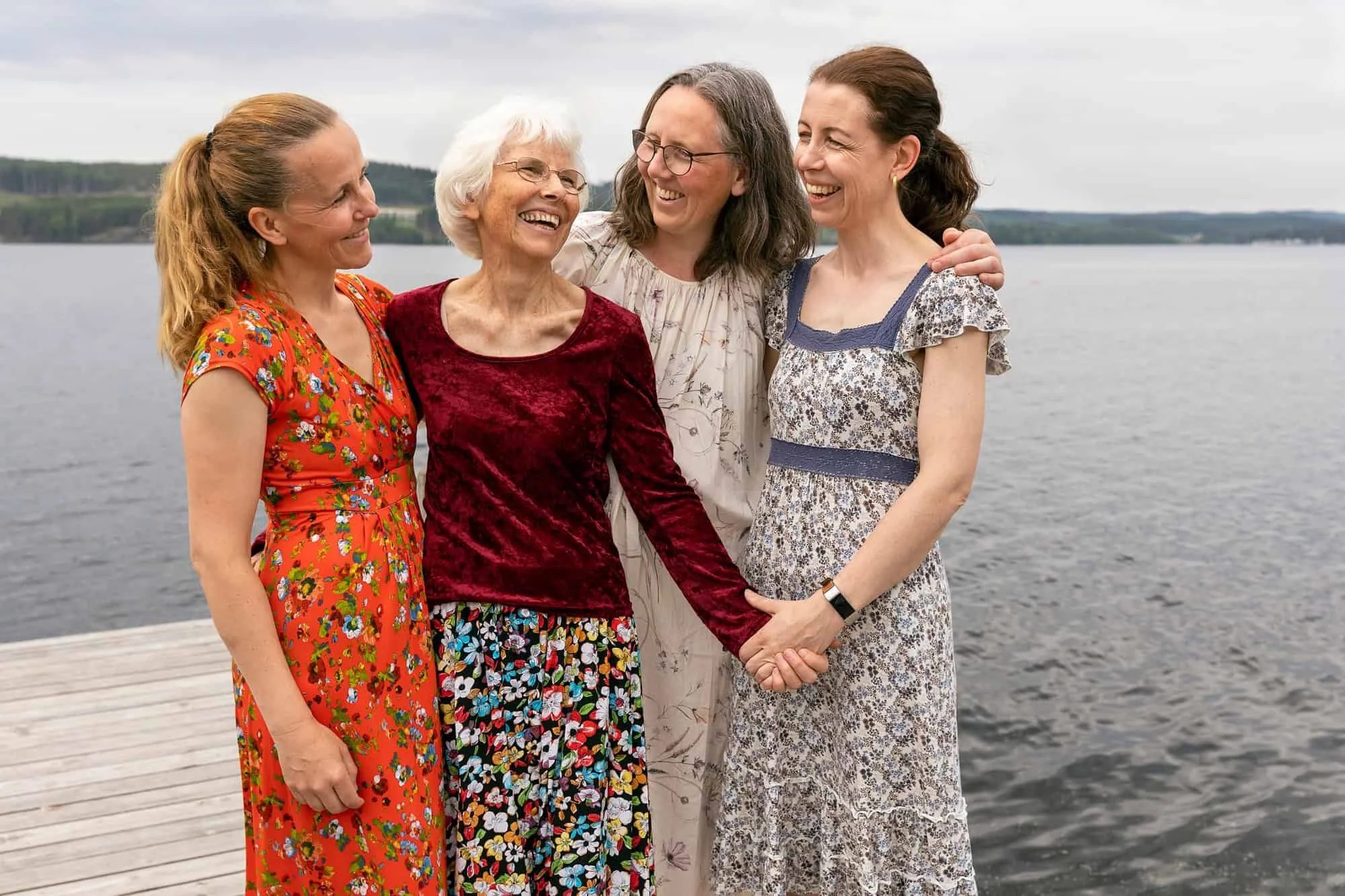 En eldre kvinne ler mellom sine voksne døtre, alle er kledd i blomstrete sommerkjoler