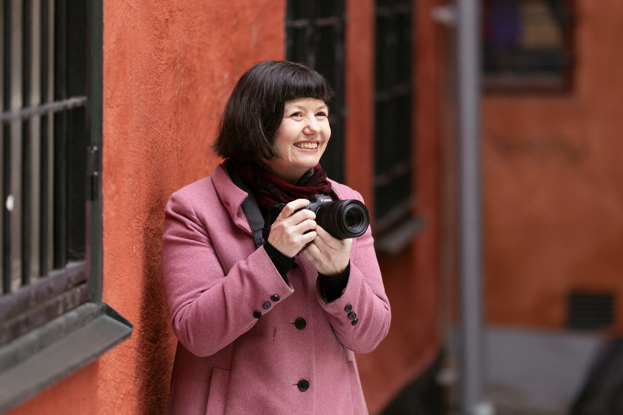 Smilende fotograf i rosa kåpe mot en rød murvegg