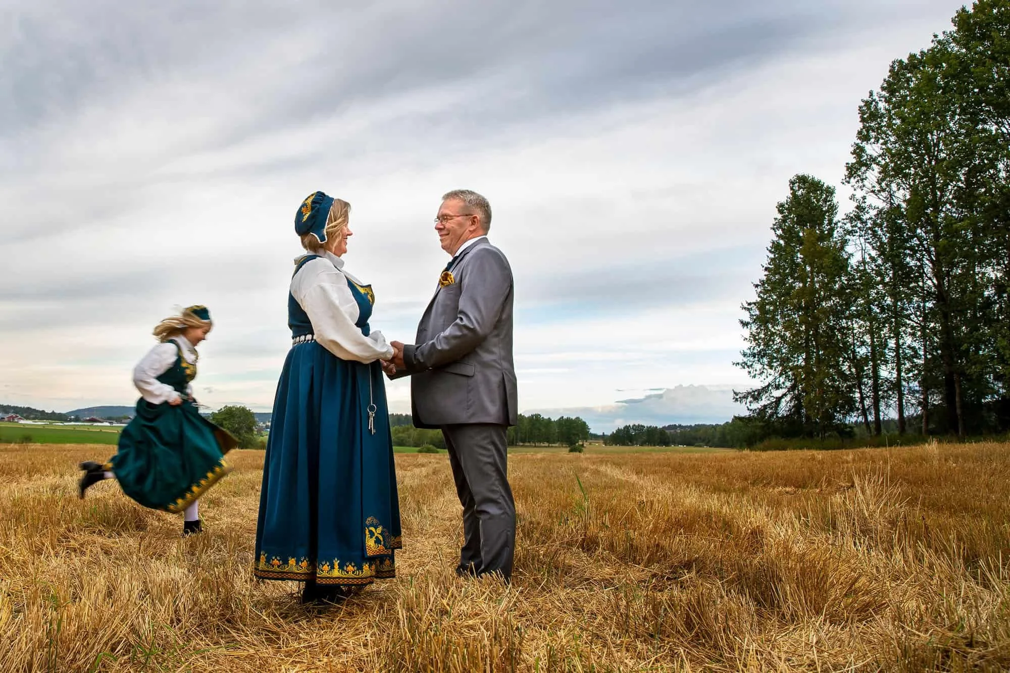 Bunadskledd jente løper i bakgrunnen av foreldrene som holder hender på en gul åker