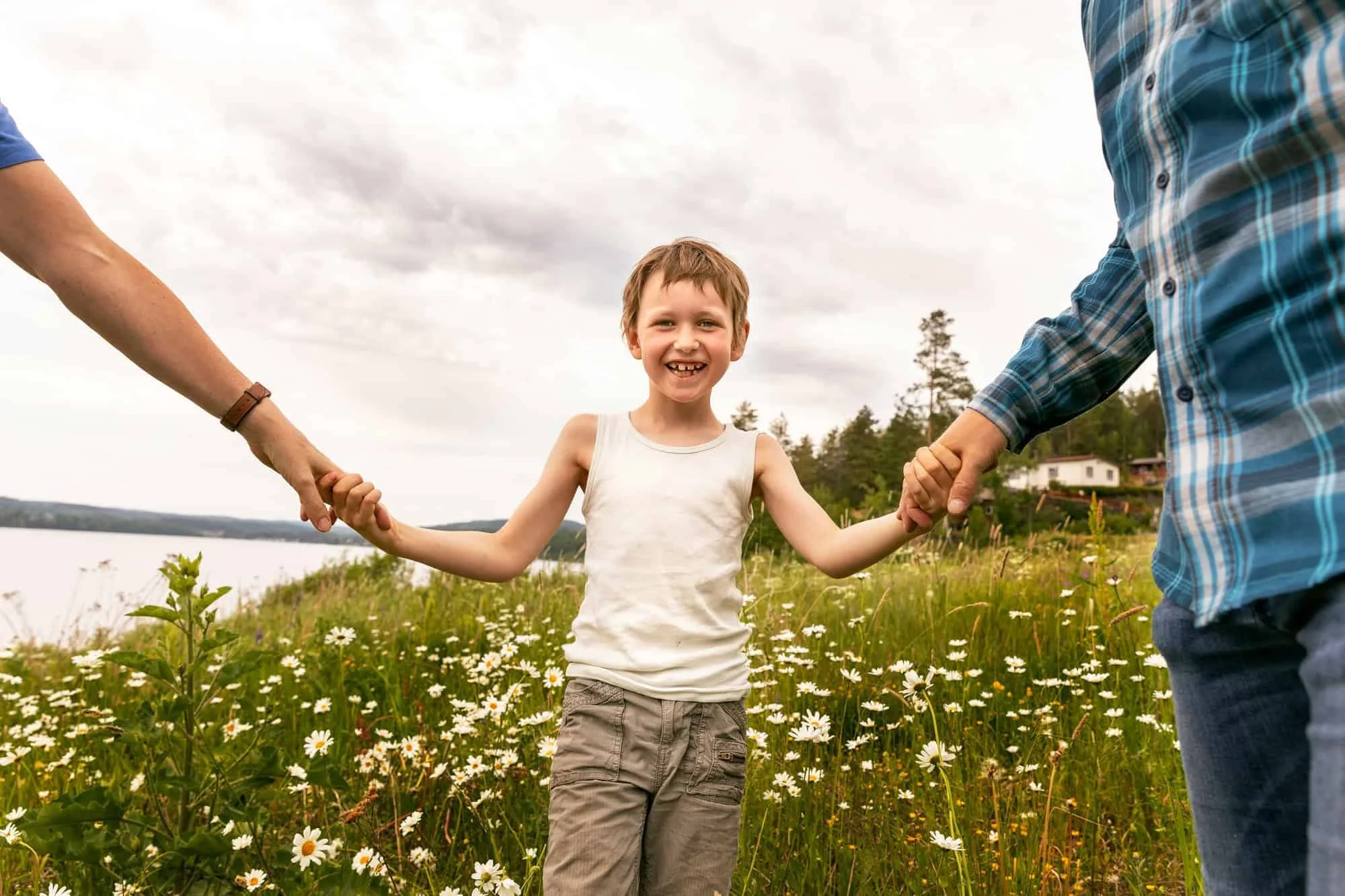 En ung gutt står i ei blomstereng og holder hånda til foreldrene sine