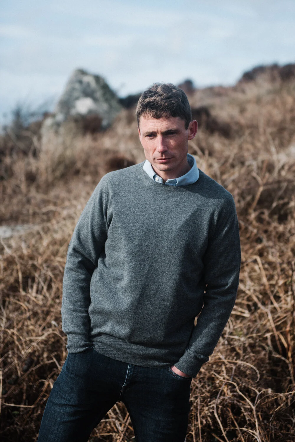 Model Matt Sharman wears a grey jumper, blue shirt and blue jeans standing on a Cornish hill for Tom Lane