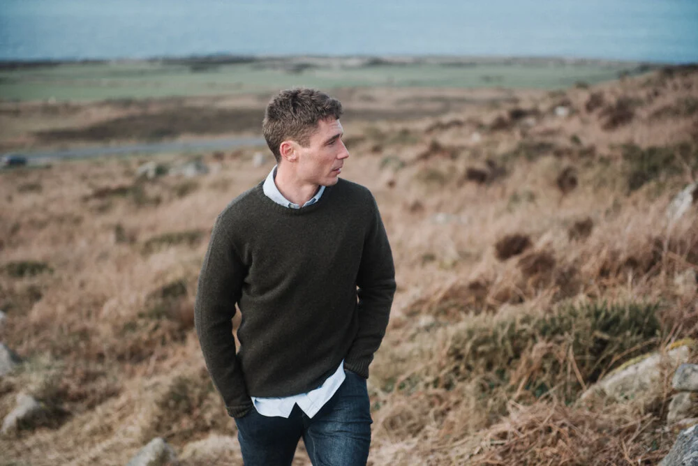 Model Matt Sharman wears a green jumper, blue shirt and blue jeans standing on a Cornish hill for Tom Lane