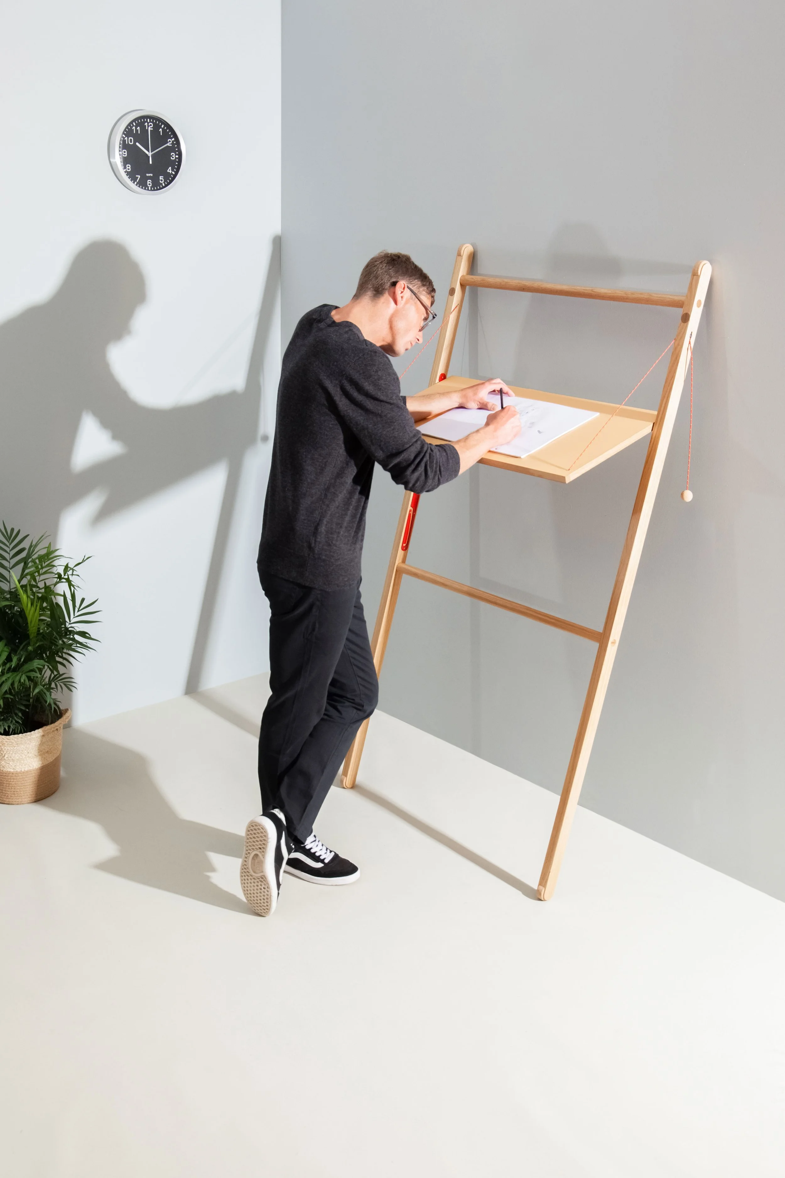 Model Matt Sharman stands at a mobile desk writing with a pencil on a pad, wearing glasses, a black jumper, black jeans and Vans trainers for Leandesk  
