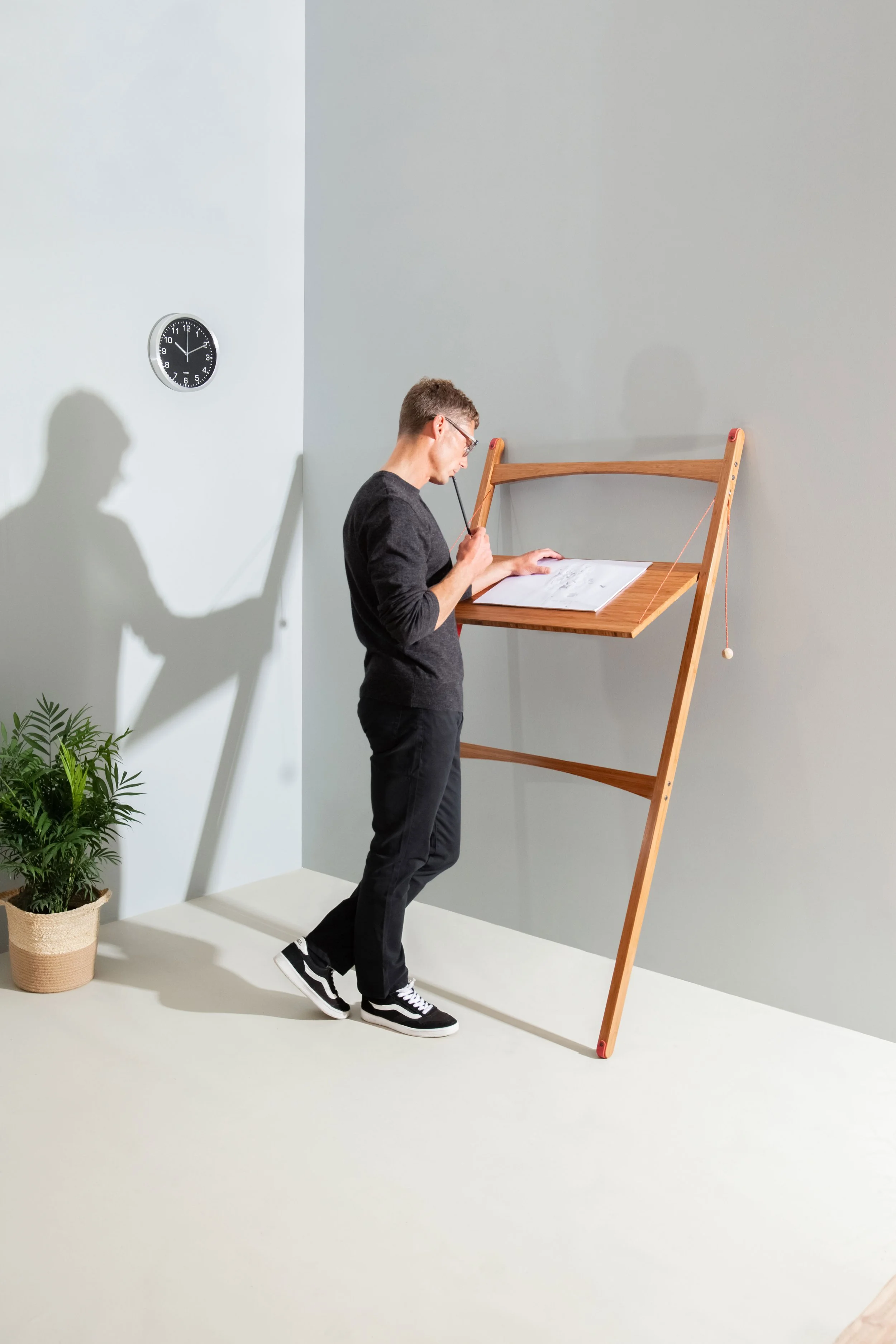 Model Matt Sharman stands at a mobile desk writing with a pencil on a pad, wearing glasses, a black jumper, black jeans and Vans trainers for Leandesk  
