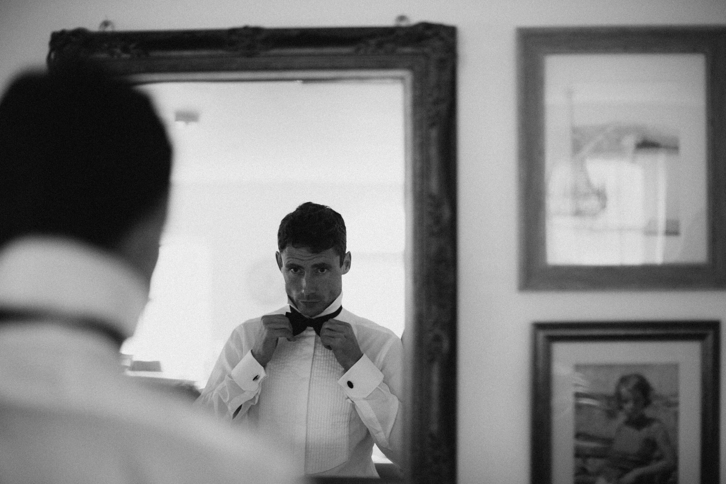 Male model Matt Sharman looks at his reflection in the mirror wearing a white shirt and black bow tie for Fern Godfrey Weddings