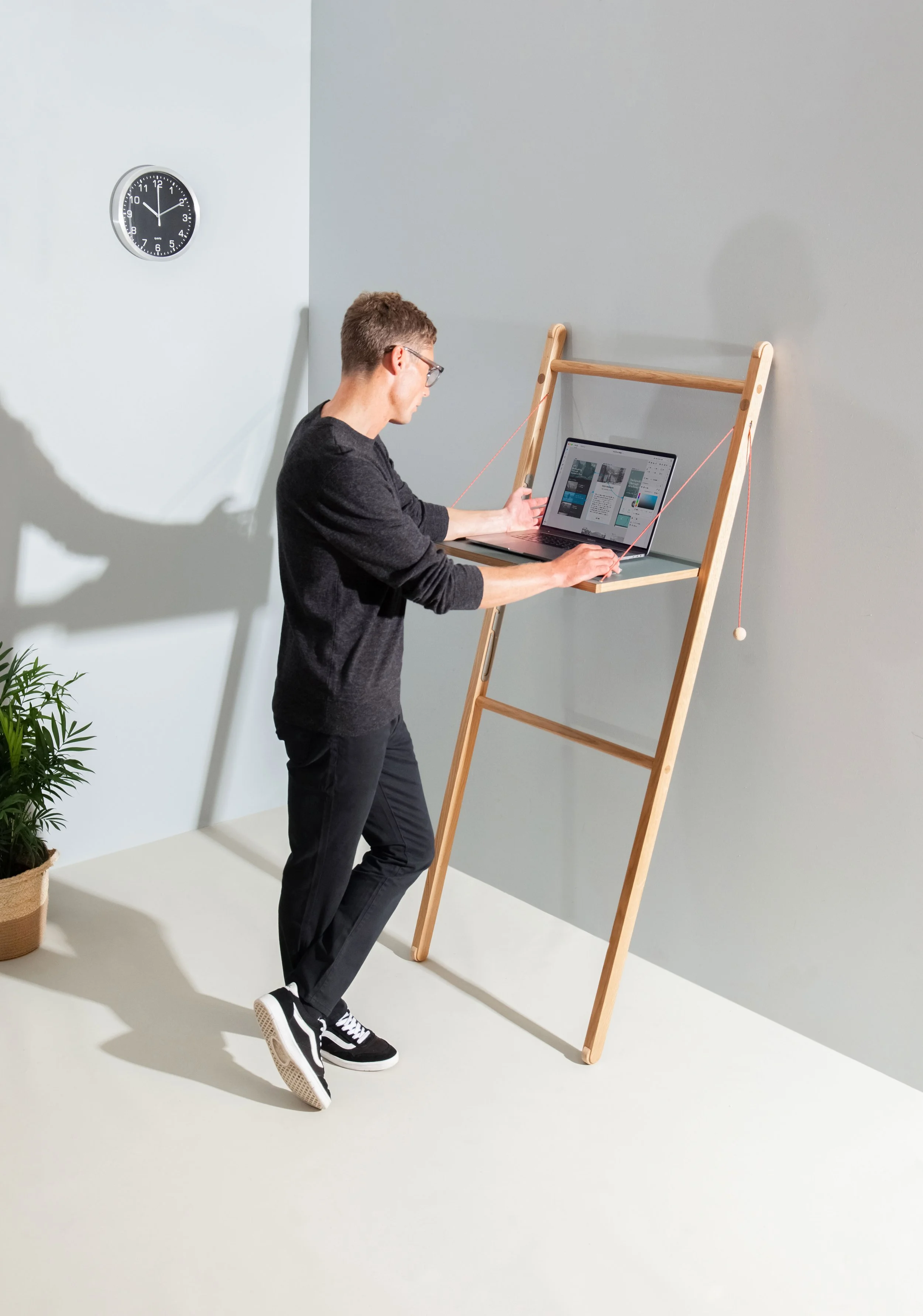 Model Matt Sharman stands at a mobile desk working on a laptop wearing glasses, a black jumper, black jeans and Vans trainers for Leandesk  