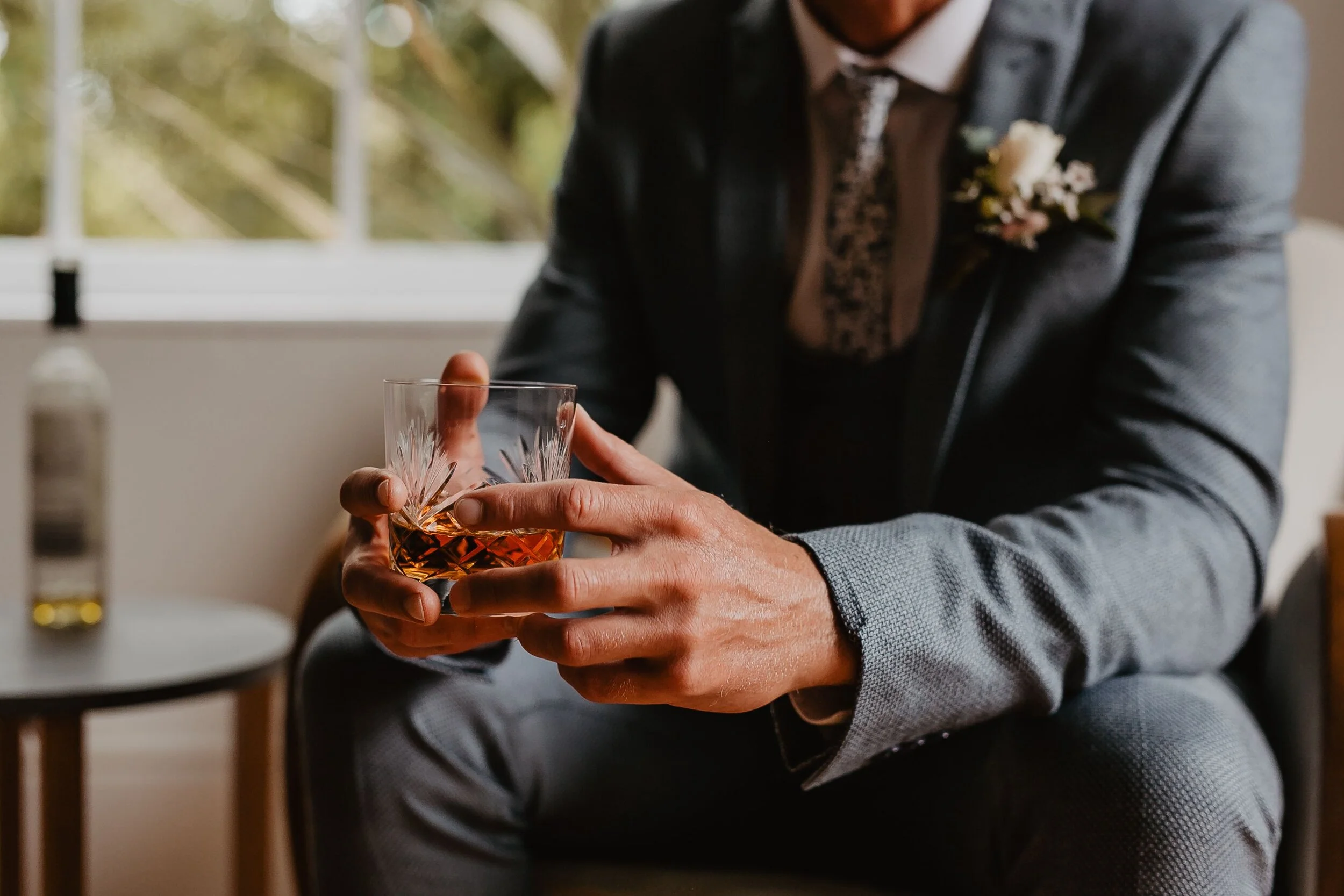 Model Matt Sharman wears a grey suit with waistcoat, white shirt and flower design tie holding a glass of whisky in a bridal shoot at Treseren House in Cornwall