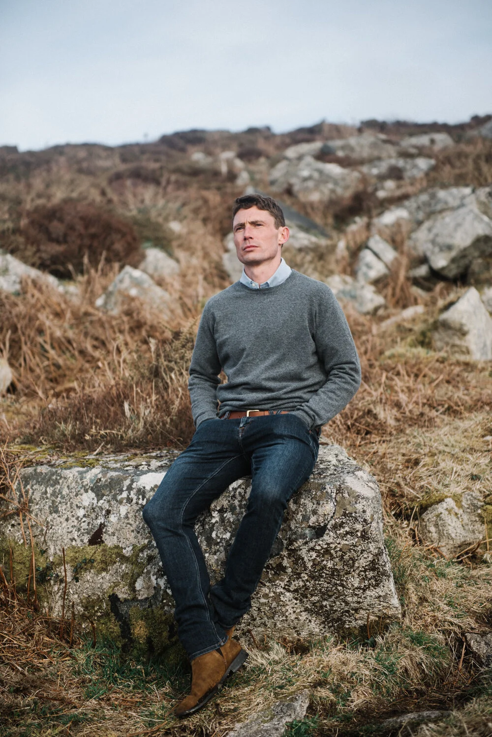 Model Matt Sharman wears a grey jumper, blue shirt and blue jeans sitting on a Cornish hill for Tom Lane