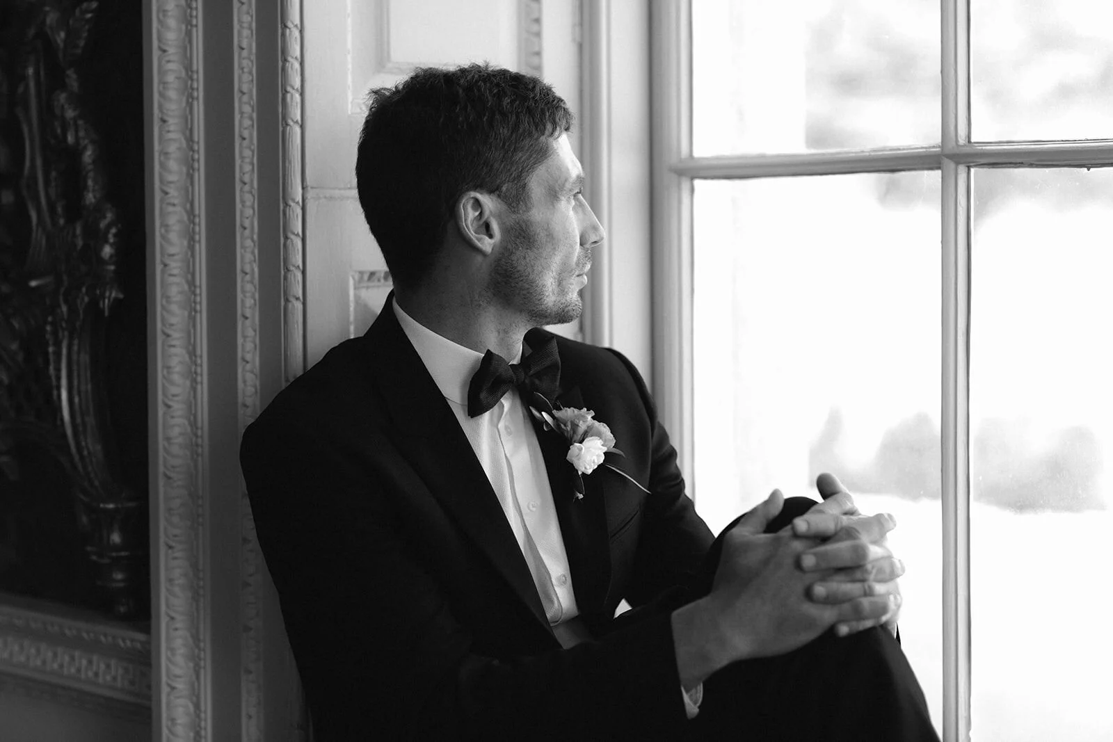 A black and white photograph of Male model Matt Sharman wearing a tuxedo with bow tie looking out of a window at luxury hire venue St Giles House in Dorset   