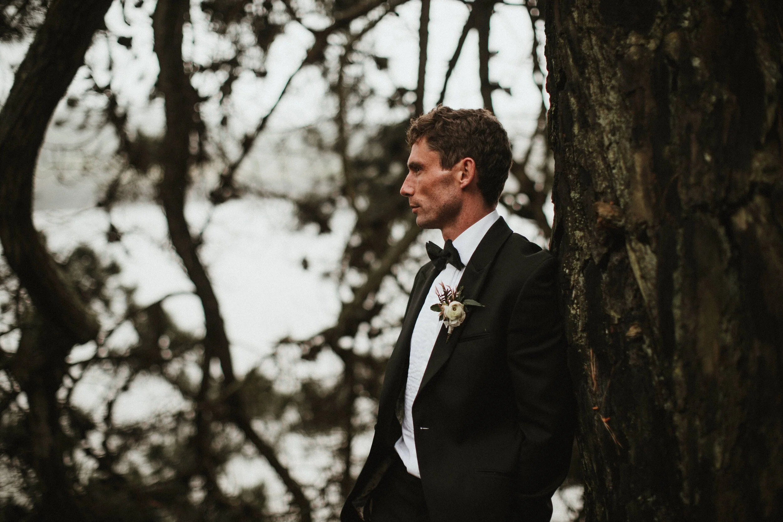 Male model Matt Sharman leans against a tree wearing a black tuxedo, white shirt and black bow tie in a Cornish forest for Fern Godfrey Weddings