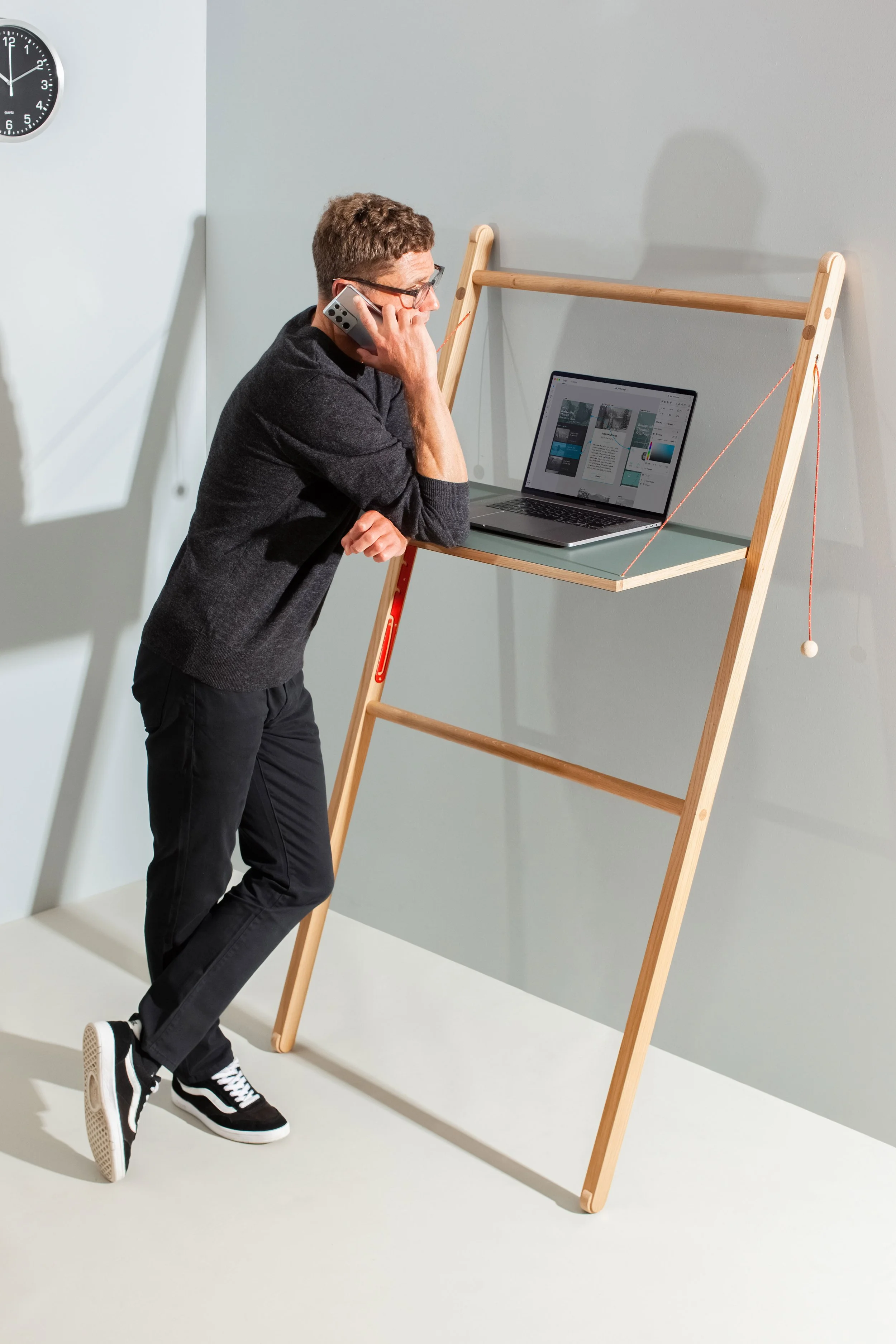 Model Matt Sharman stands at a mobile desk talking on a mobile telephone, wearing glasses, a black jumper, black jeans and Vans trainers for Leandesk  
