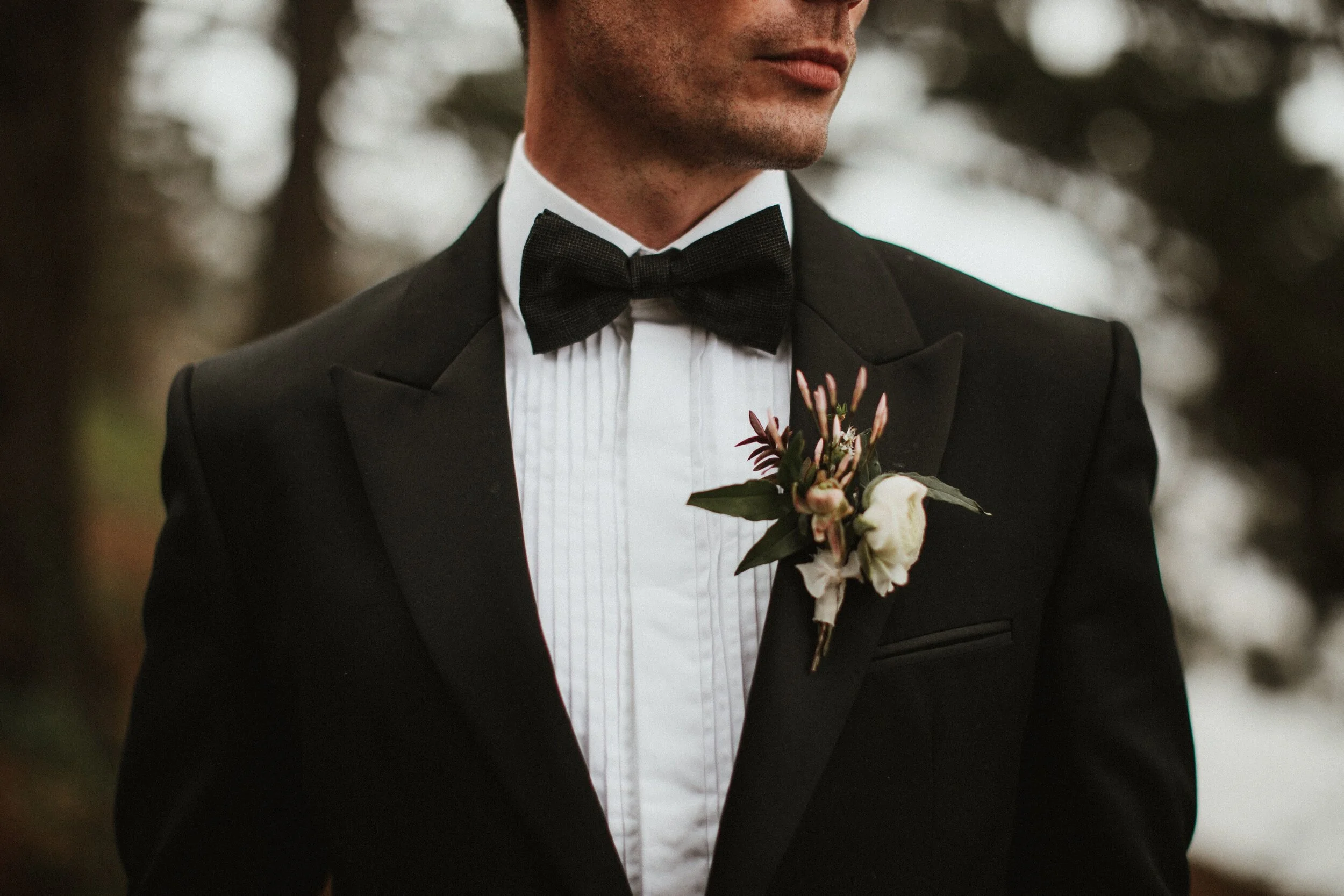 A close up shot of male model Matt Sharman wearing a black tuxedo, white shirt and black bow tie in a Cornish forest for Fern Godfrey Weddings