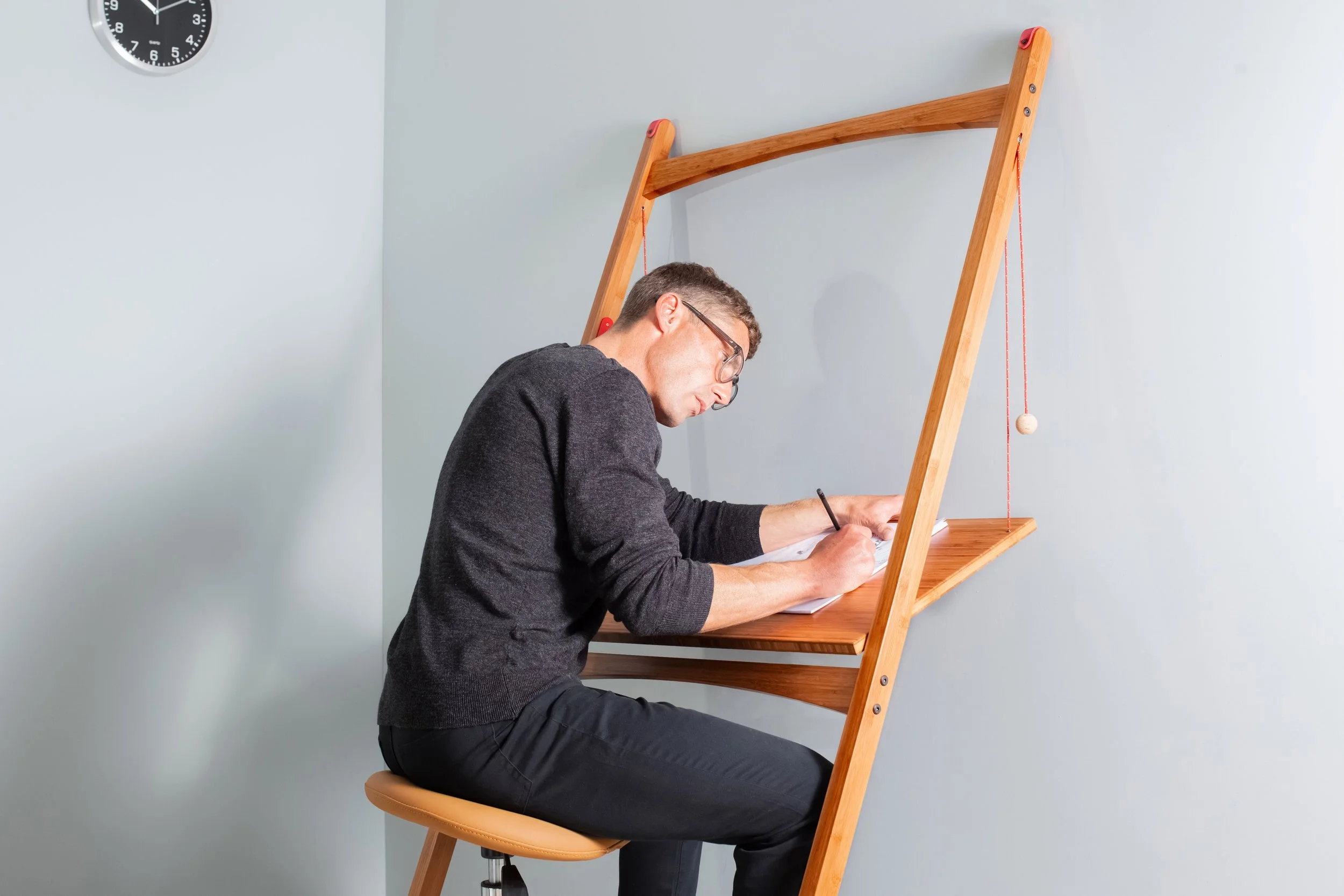 Model Matt Sharman sits at a mobile desk writing with a pencil on a pad, wearing glasses, a black jumper, black jeans and Vans trainers for Leandesk 