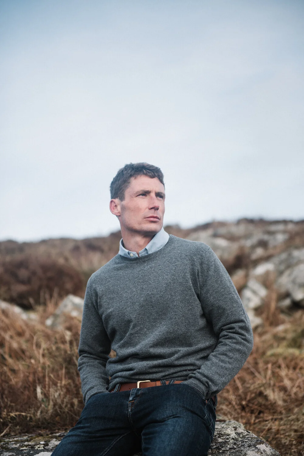 Model Matt Sharman wears a grey jumper, blue shirt and blue jeans sitting on a Cornish hill for Tom Lane