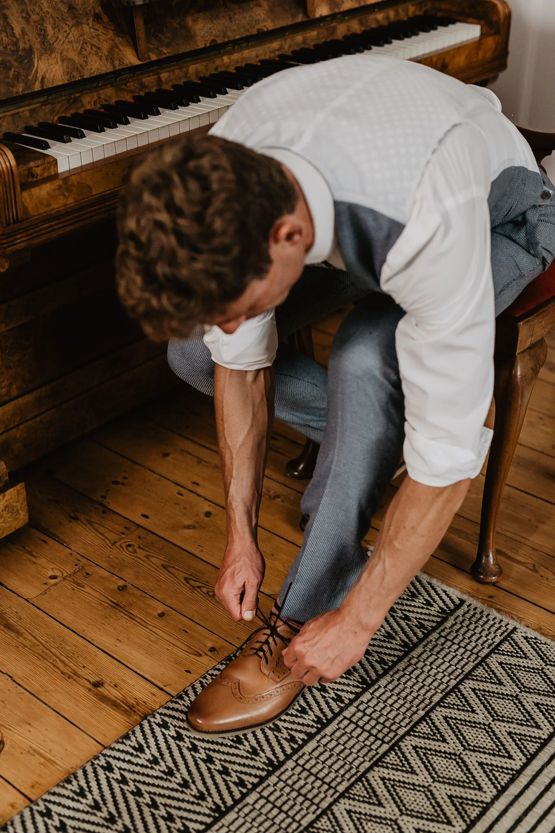 Model Matt Sharman wears a grey suit with waistcoat, white shirt and flower design tie lacing a shoe lace of brown shoes in a bridal shoot at Treseren House in Cornwall