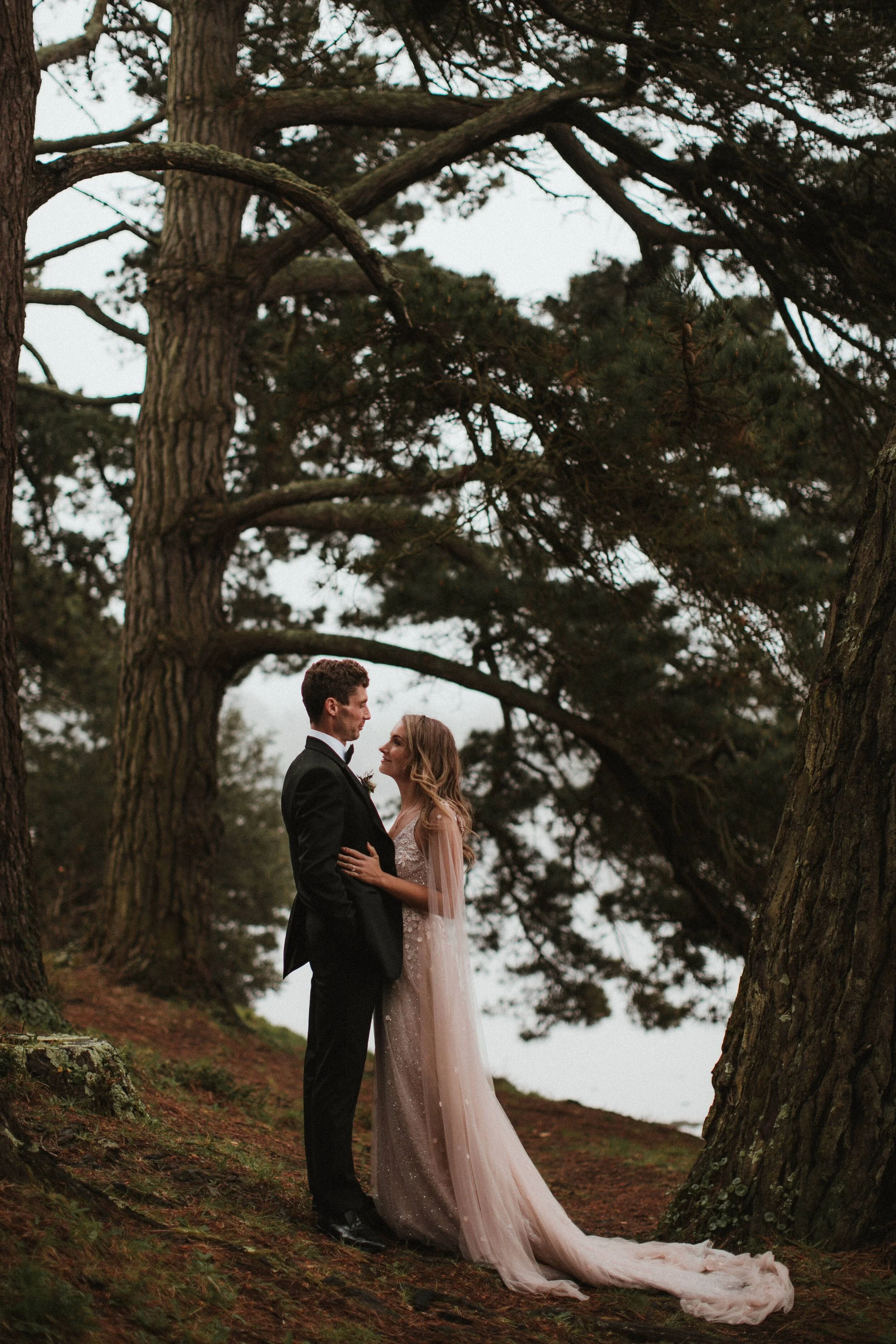Male model Matt Sharman holds female model Lara wearing a black tuxedo, white shirt and black bow tie in a Cornish forest for Fern Godfrey Weddings