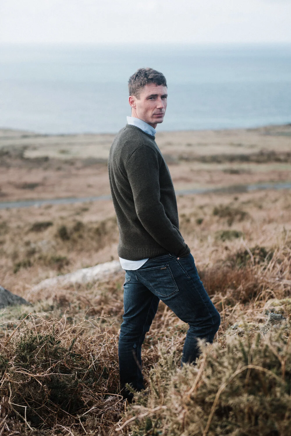 Model Matt Sharman wears a green jumper, blue shirt and blue jeans standing on a Cornish hill for Tom Lane 