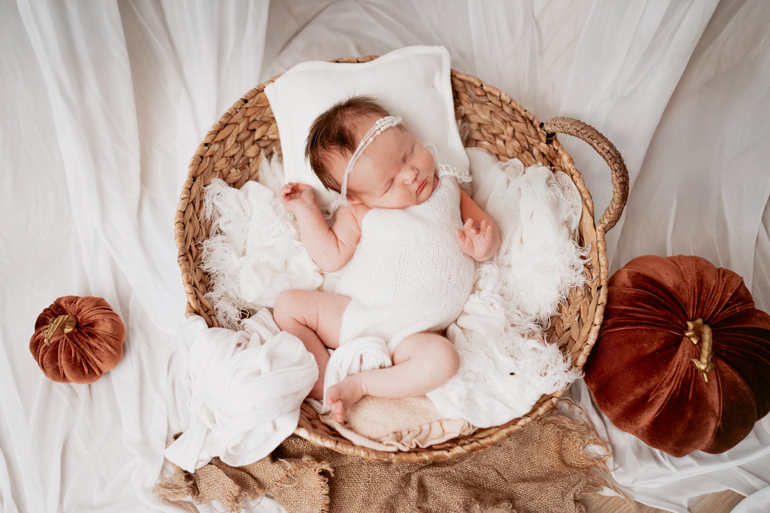 Newborn baby peacefully sleeping in a cozy, rustic autumn-themed setup. The baby is wrapped in a soft beige knitted blanket, wearing a fluffy bear-ear bonnet, and holding a tiny heart-shaped pillow. The scene is adorned with warm-toned dried leaves