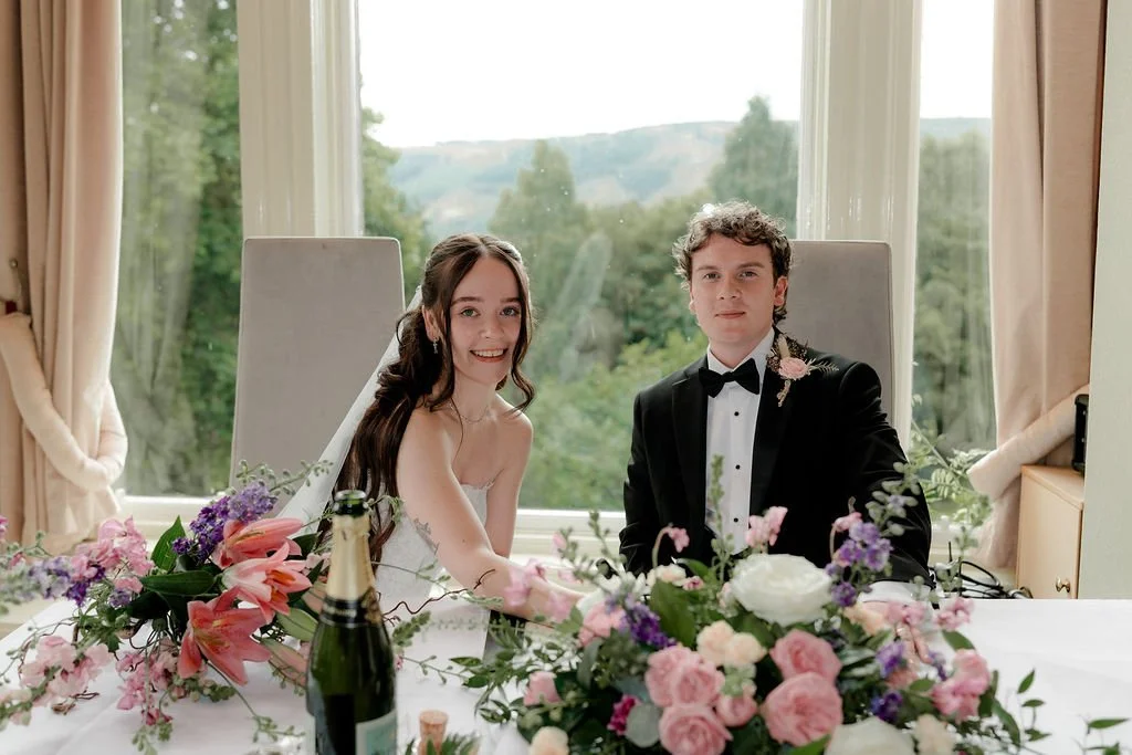 Bride and groom seated at their wedding table behind colourful pink and purple flowers, with woodland views through the windows at Atholl Palace Hotel.