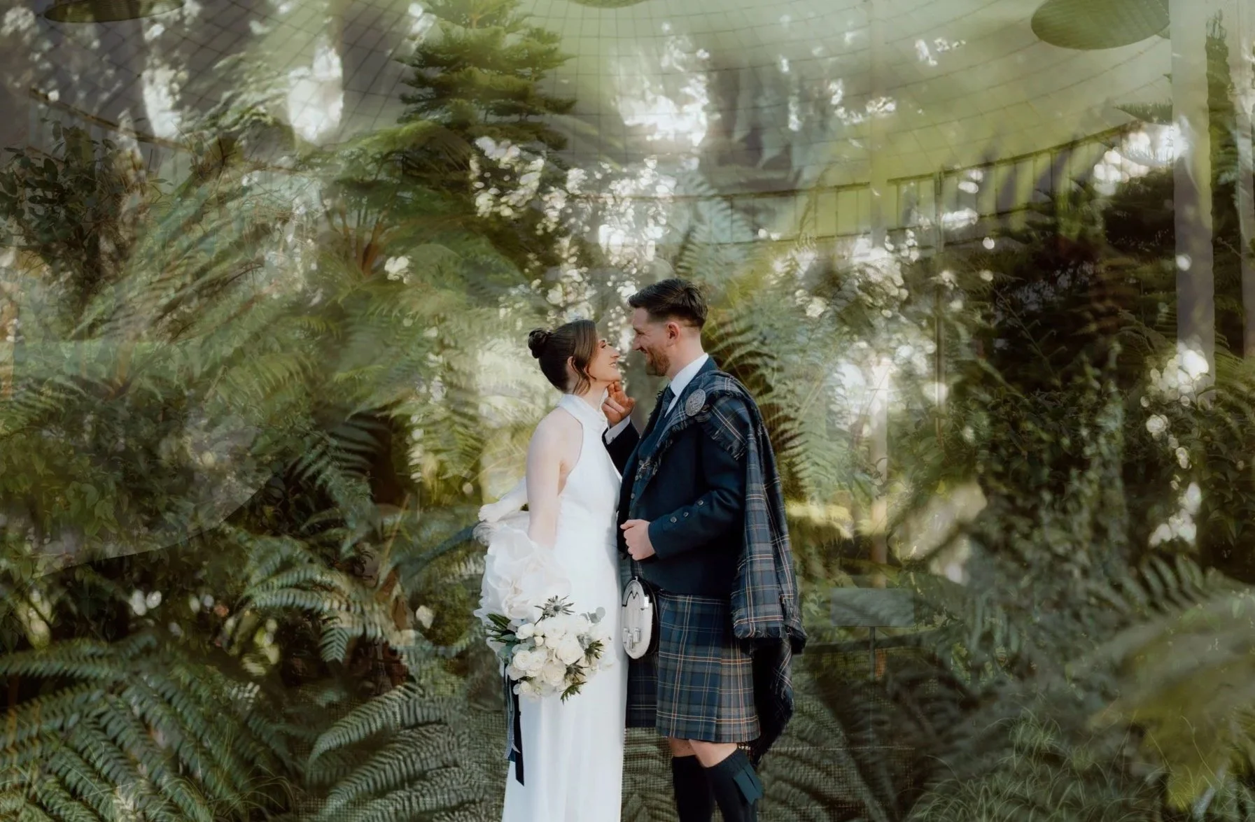 A bride and groom sharing a romantic moment in a lush, green outdoor setting with ferns in the background, the bride wearing a white dress and the groom in a dark suit.