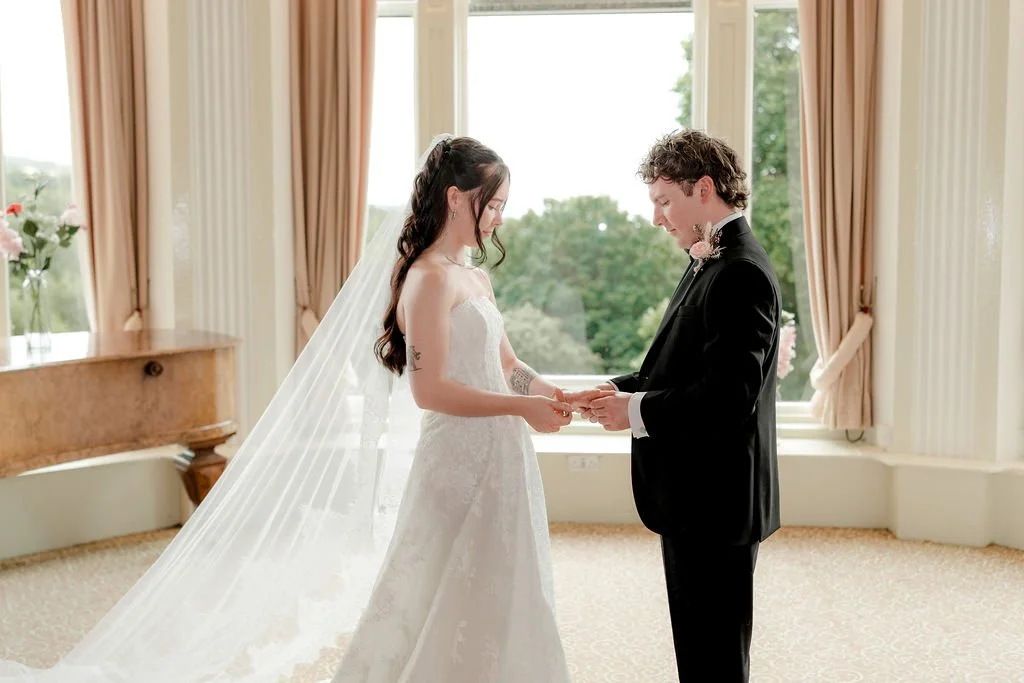 Bride and groom holding hands and exchanging rings in front of tall, light-filled windows during their Atholl Palace wedding ceremony.