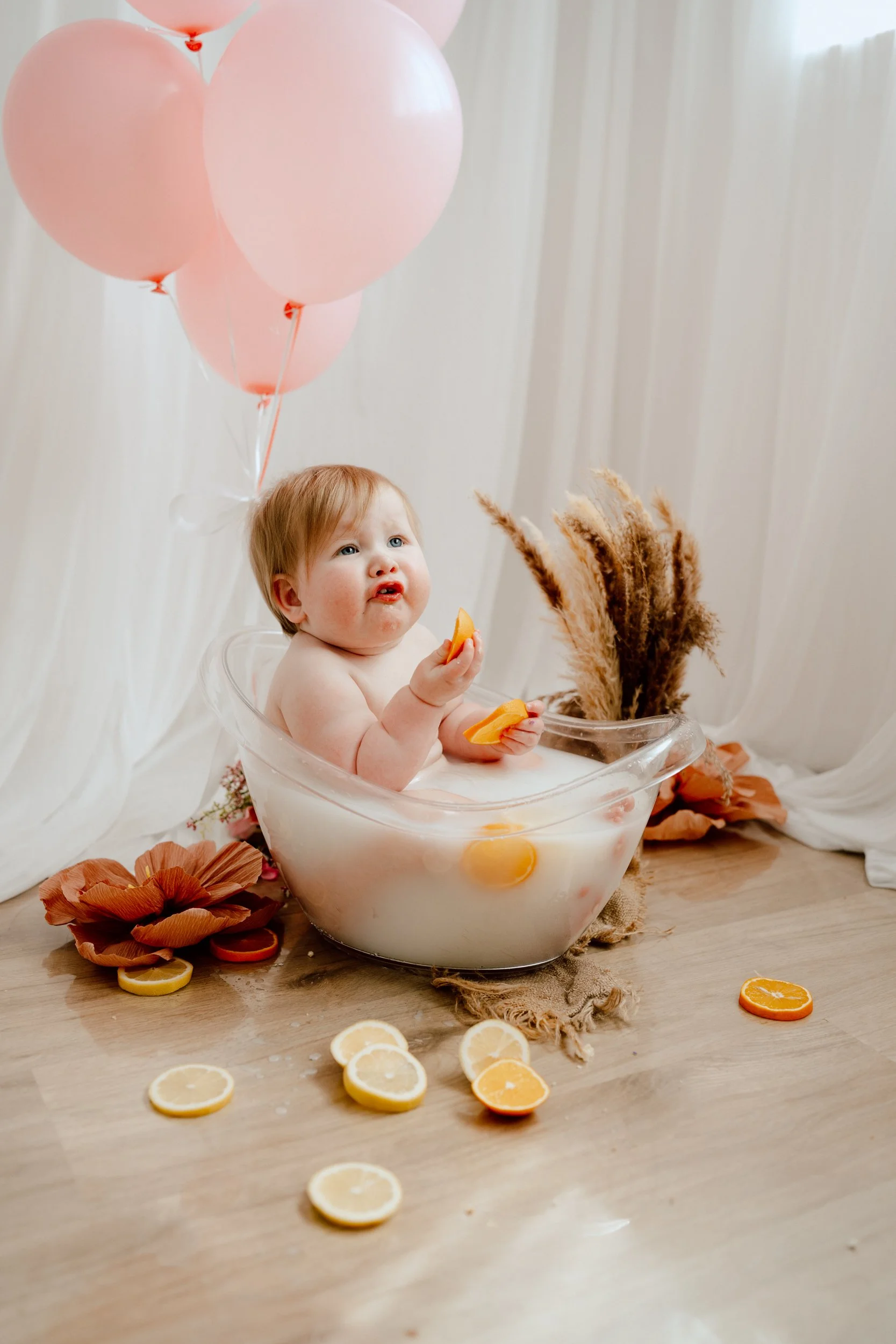 Toddler sitting in a clear milky bath holding citrus fruit with at boho setup durig their cake smash and splash photoshoot in studio in perthshsire.