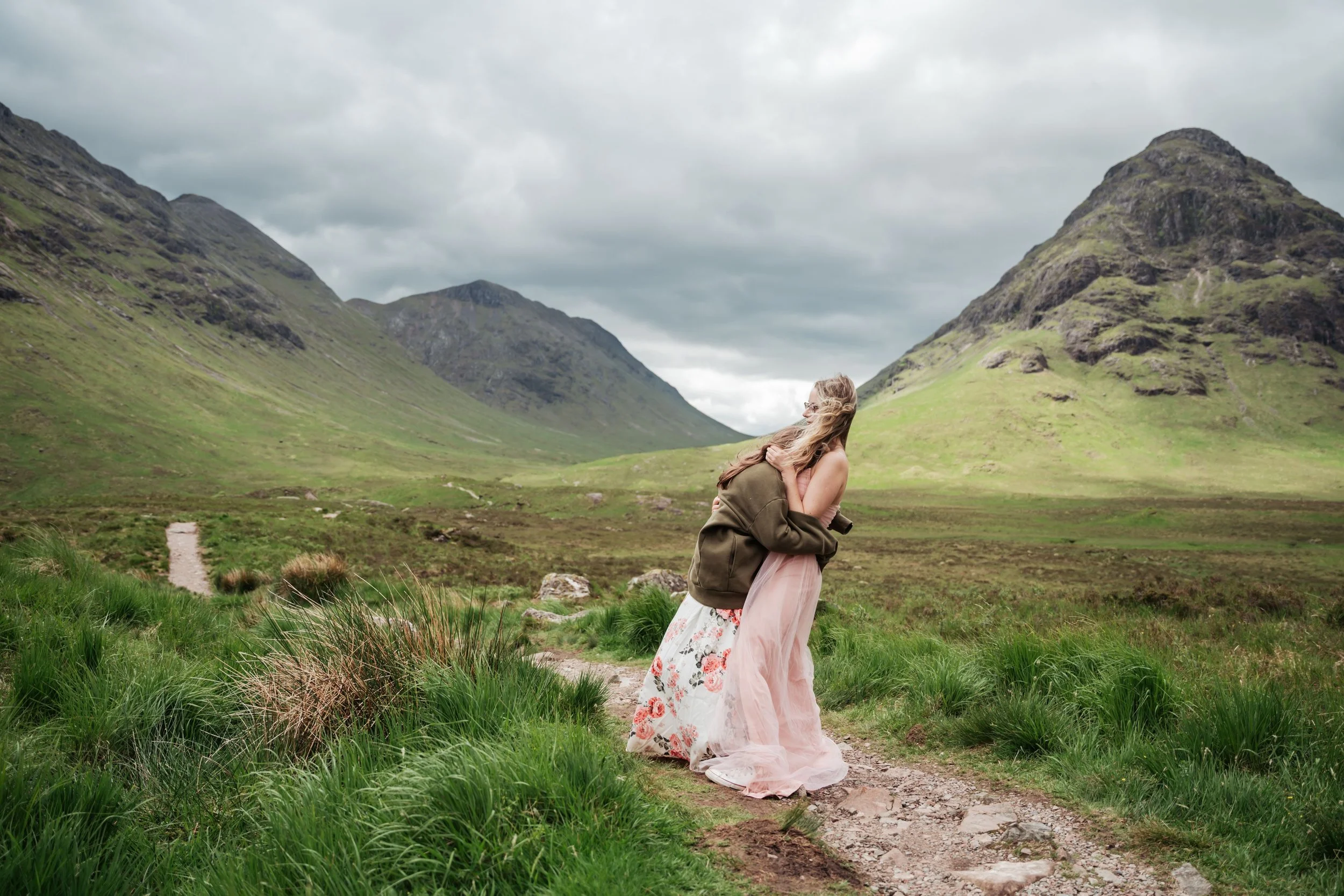 A woman in a pink dress and an olive green jacket hugging another woman on a dirt trail in a green valley surrounded by Three sisters mountains under cloudy sky.