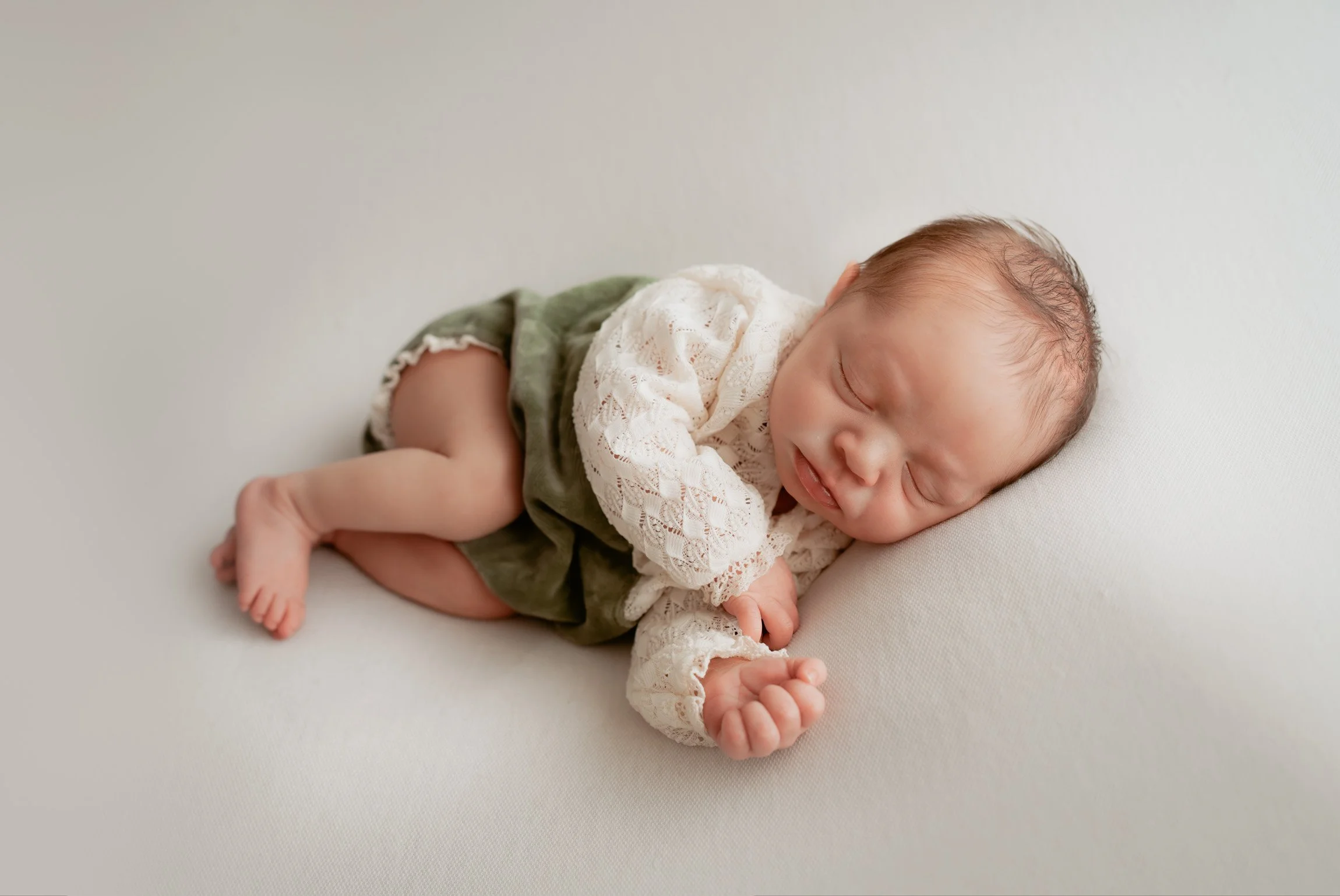 Newborn baby sleeping with her legs naturally curled, wearing a cream lace top and muted green outfit on a neutral studio backdrop.