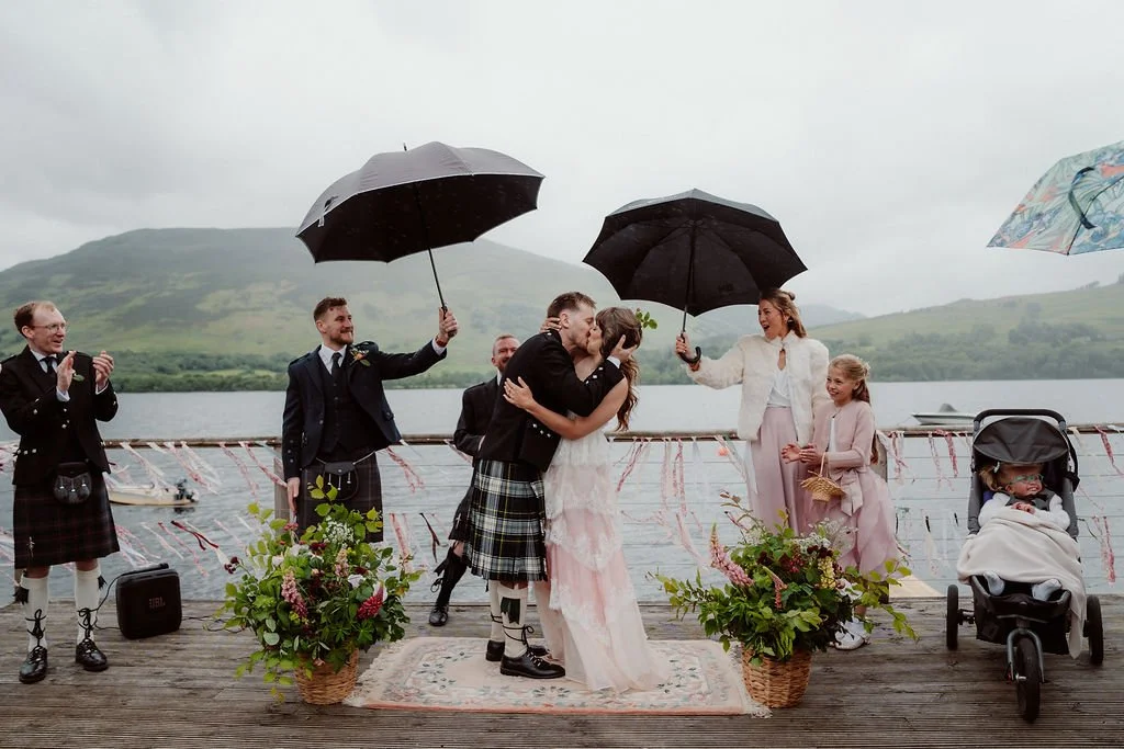 Newlywed couple kissing in the rain on a wooden pier beside a Scottish loch, surrounded by family and friends holding umbrellas at Loch Earn photographed by Patrice Little Photography.